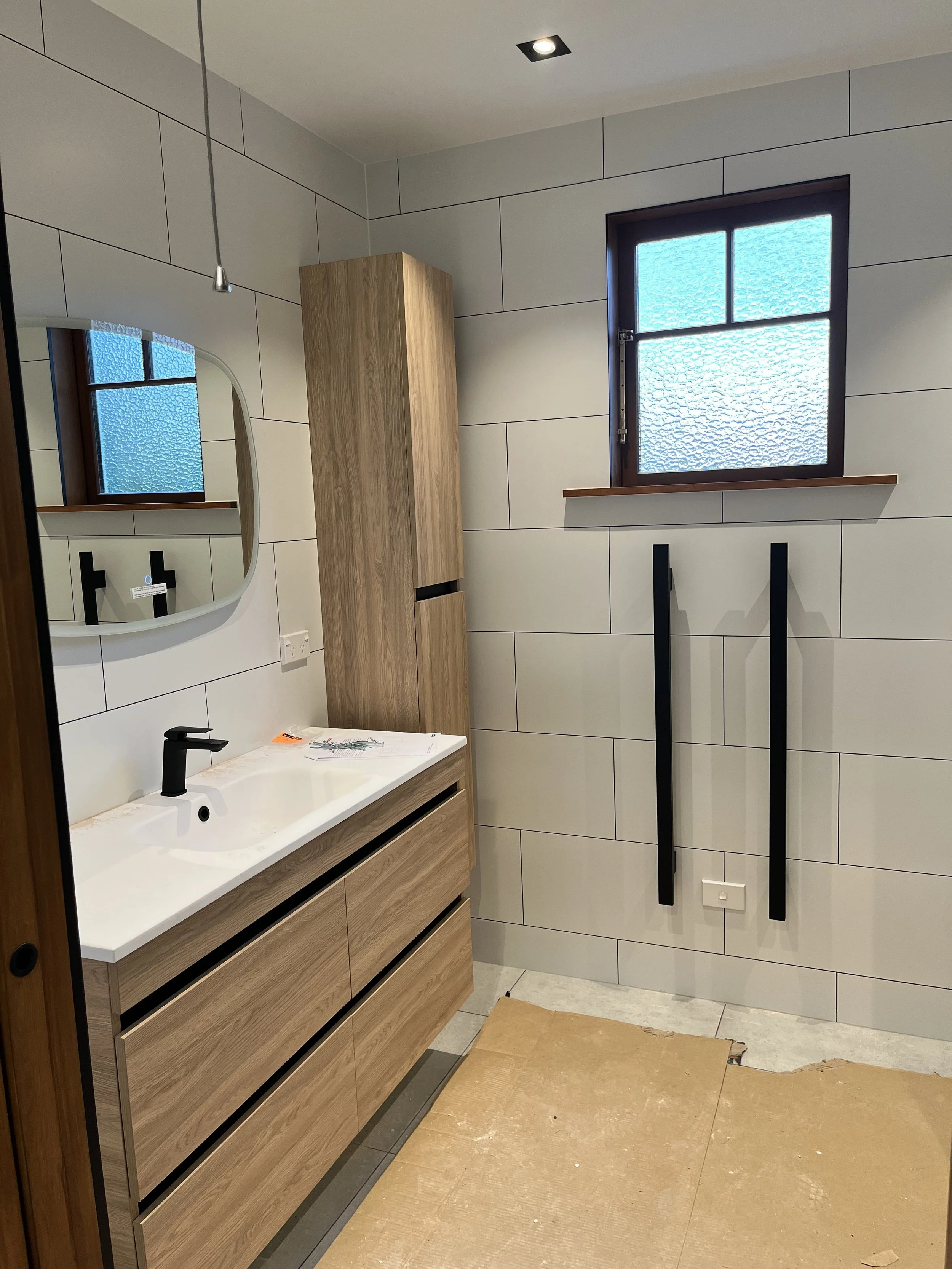 A modern bathroom with a wooden vanity and a white sink, black faucet, an oval mirror, a wooden cabinet, a textured glass window, and black towel bars on the wall.