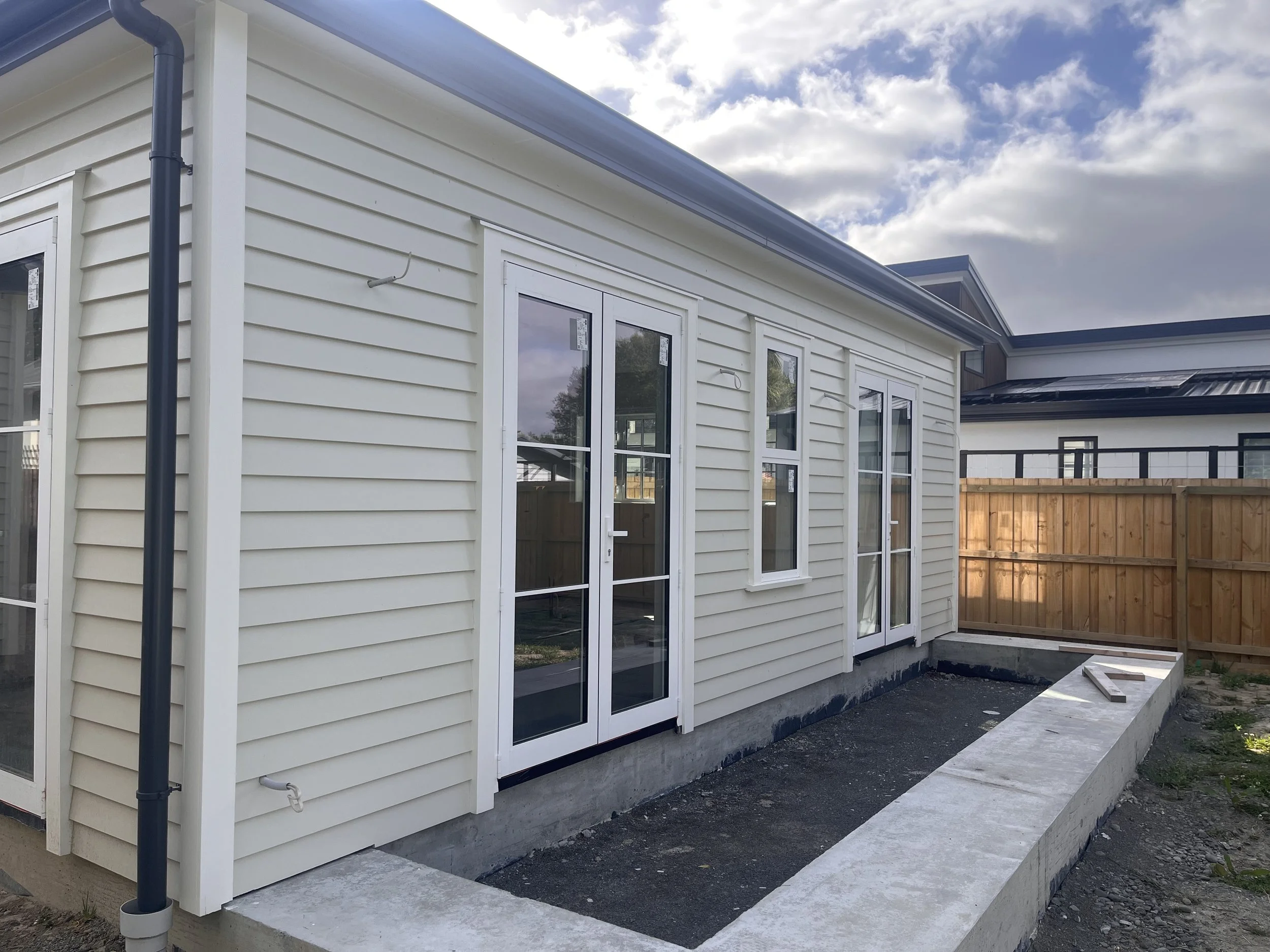 Newly constructed white house exterior with vinyl siding, glass doors, and a concrete foundation, featuring a fenced backyard with a wooden privacy fence under a partly cloudy sky.