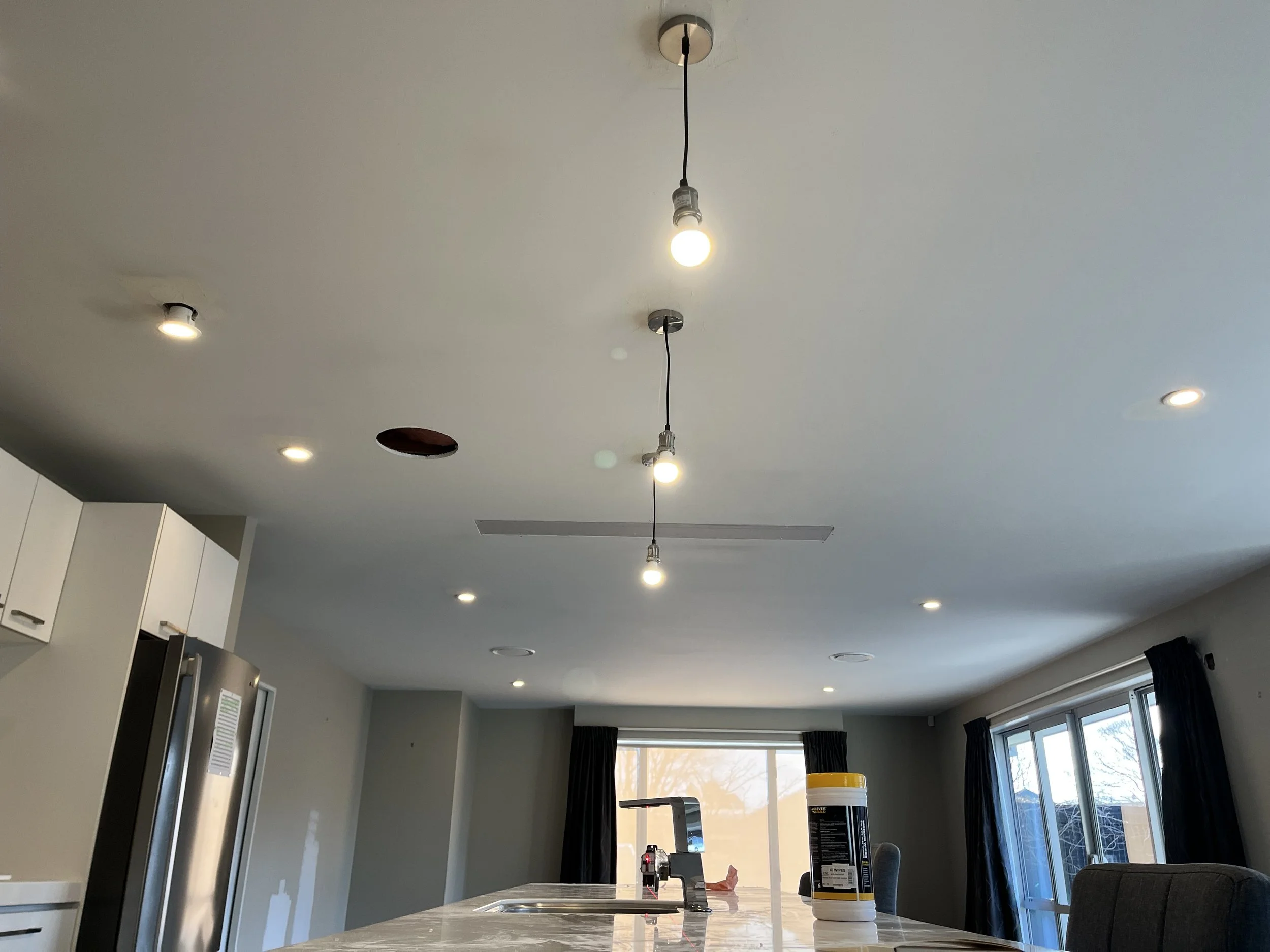 Empty kitchen with missing ceiling light fixtures and hanging light bulbs, a kitchen island with a bottle of disinfectant wipes and a sink, and large windows with black curtains.