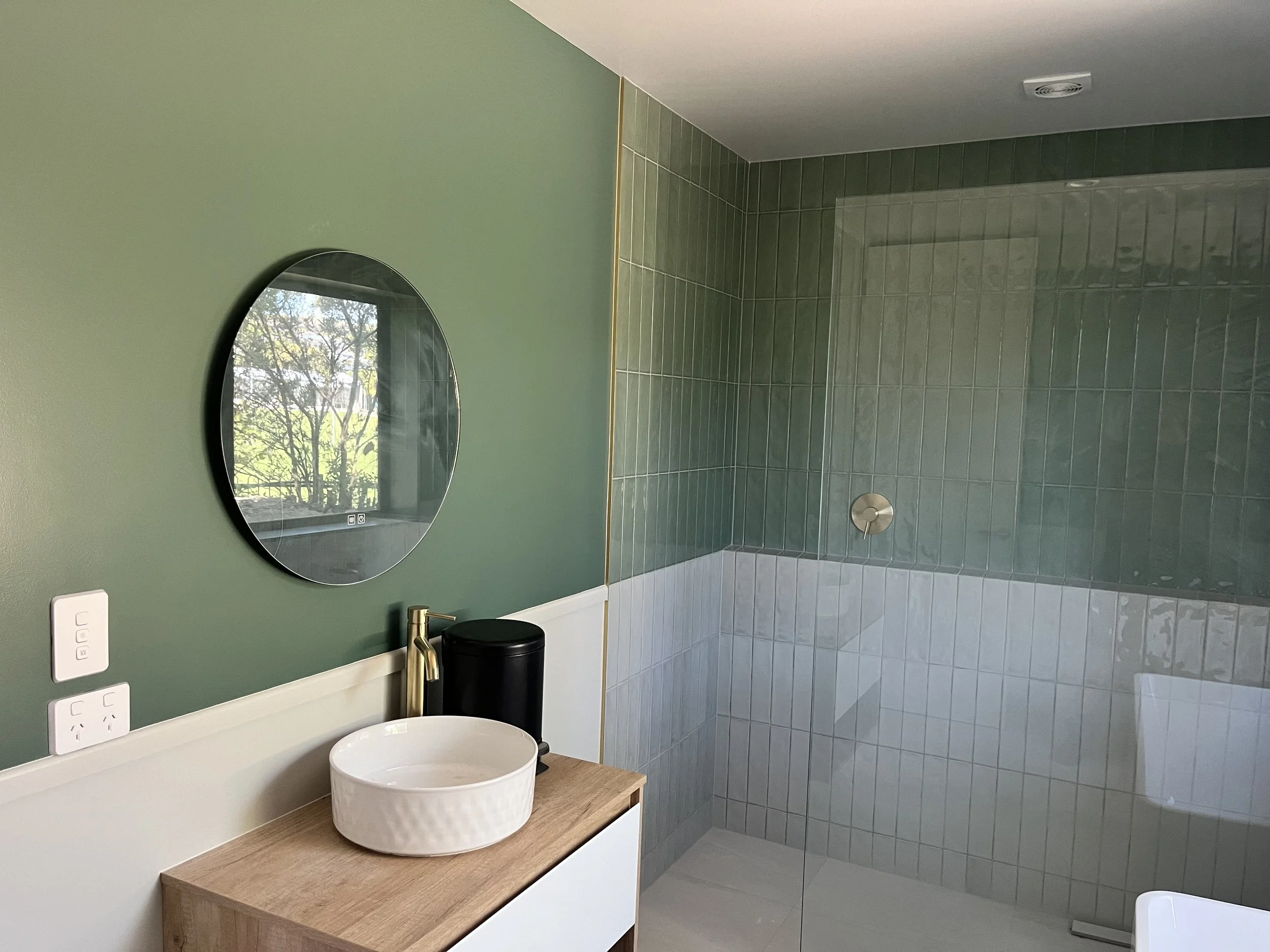 Modern bathroom with a sight view of trees outside the window, featuring a green wall, a round mirror, a wooden vanity with a white vessel sink, and a glass-enclosed walk-in shower with green and white tiles.