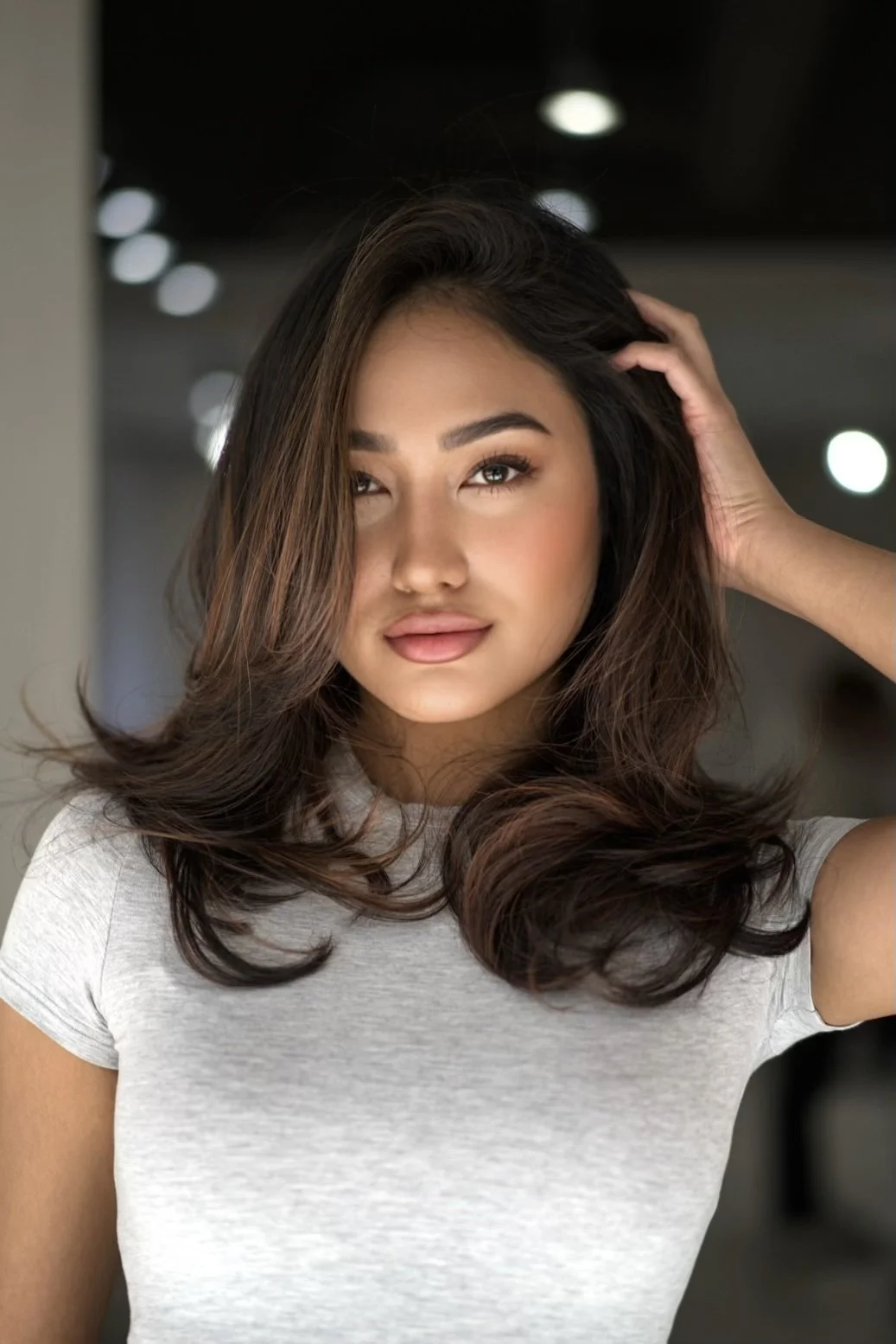 A young woman with wavy dark hair and light makeup, wearing a light gray T-shirt, posing indoors with her hand touching her hair.