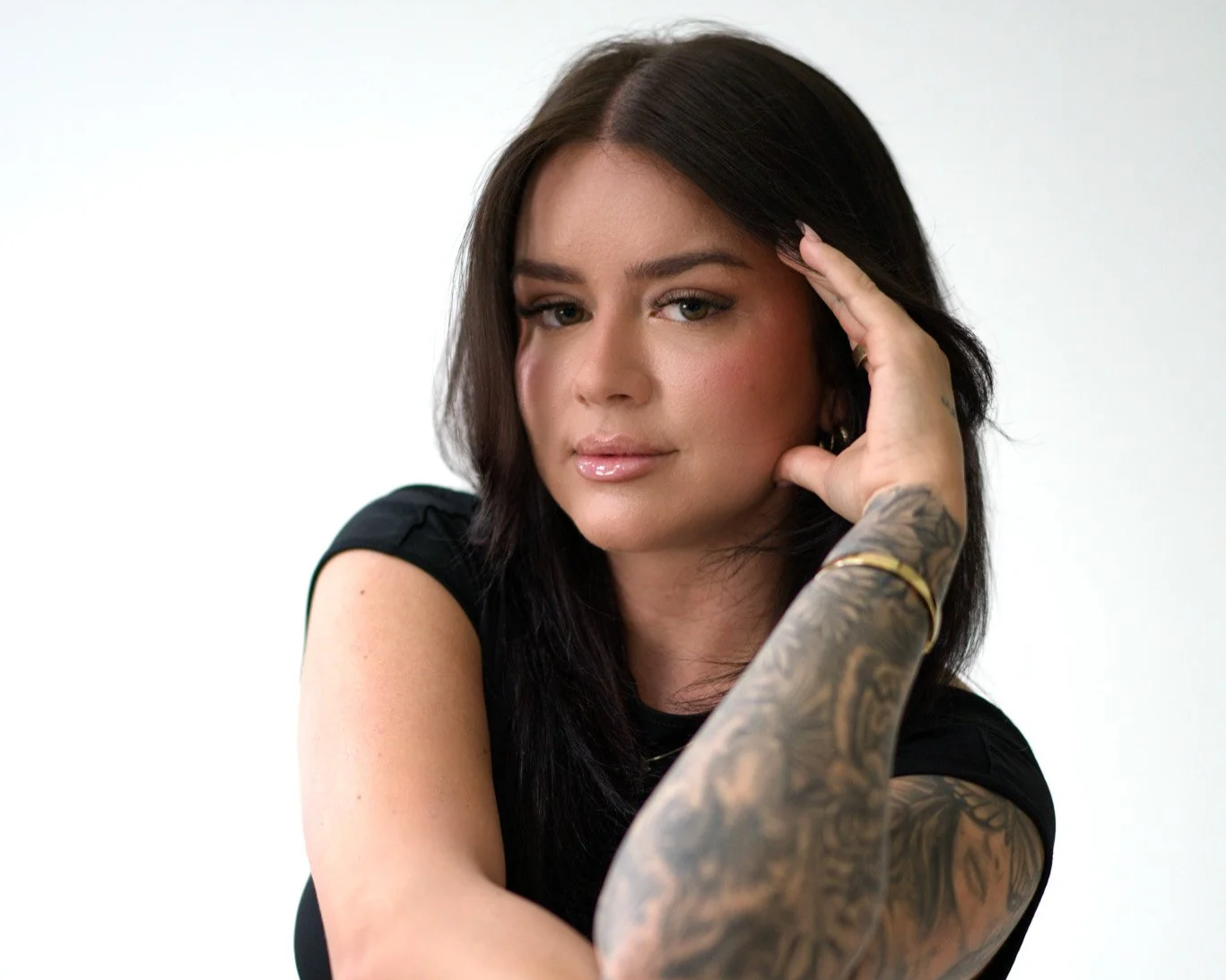 A woman with dark brown hair, light makeup, and a tattooed arm, wearing a black top, posed with her hand resting on her temple against a plain white background.
