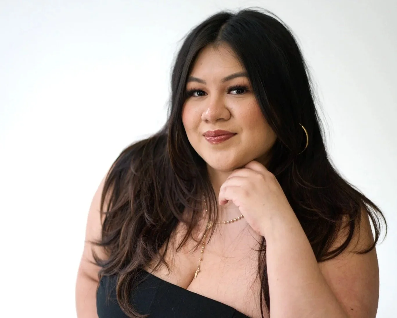 A woman with long dark hair, wearing makeup, hoop earrings, and layered necklaces, resting her chin on her hand, looking at the camera with a slight smile against a plain white background.