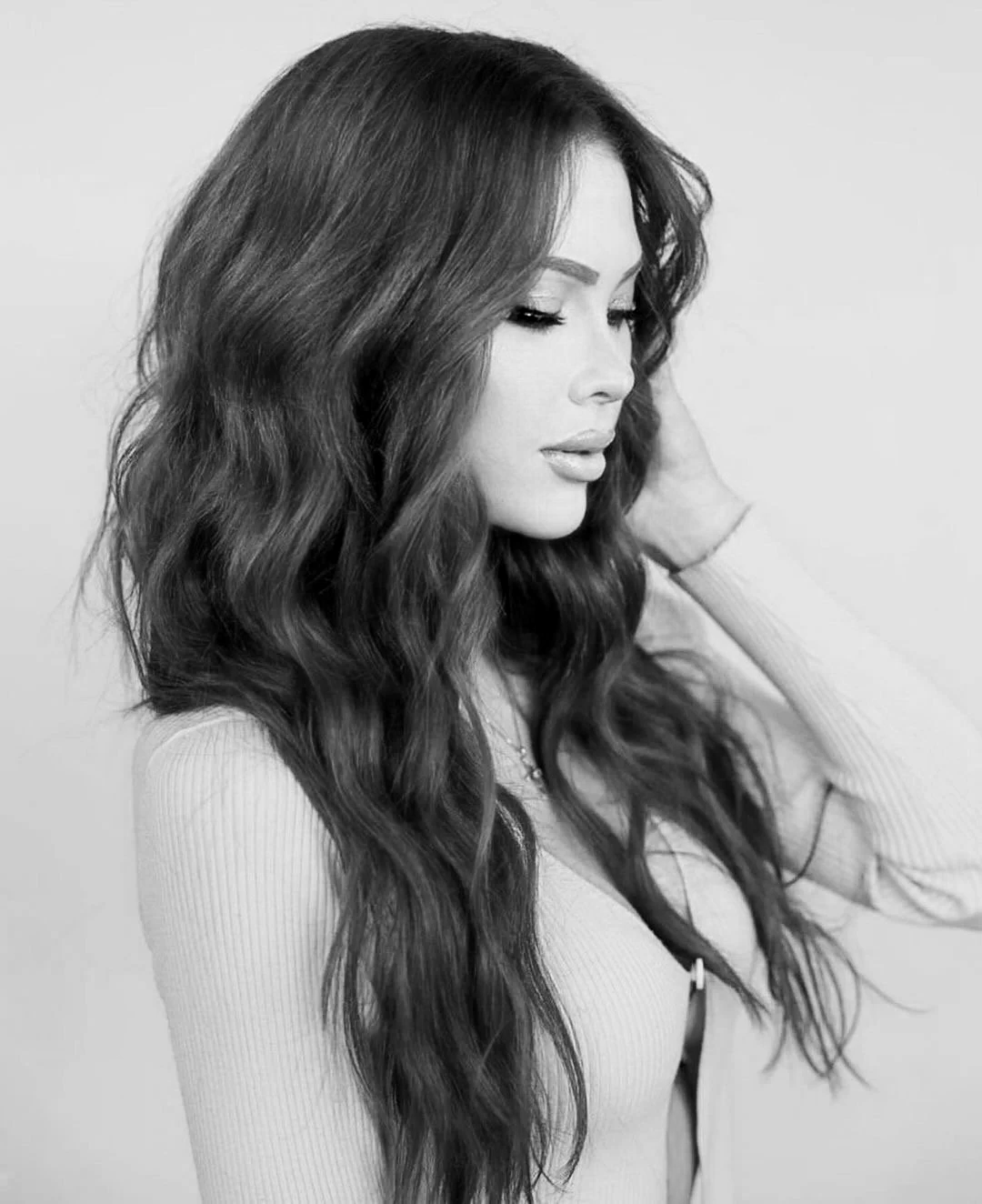 A woman with long wavy hair looking down, touching her hair with her hand, in black and white.