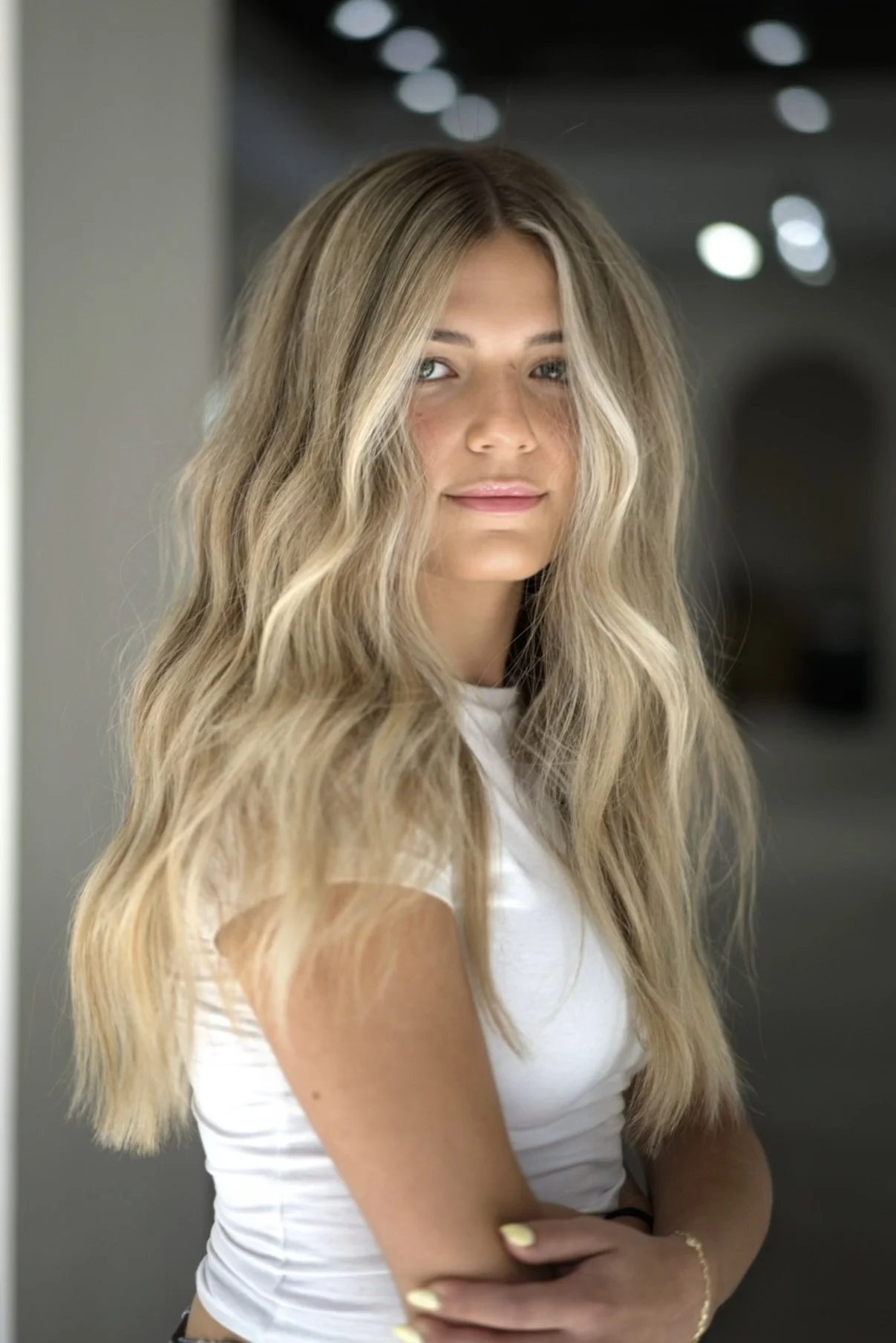 A young woman with long, wavy blonde hair wearing a white t-shirt, standing indoors with dark background and blurred lights.