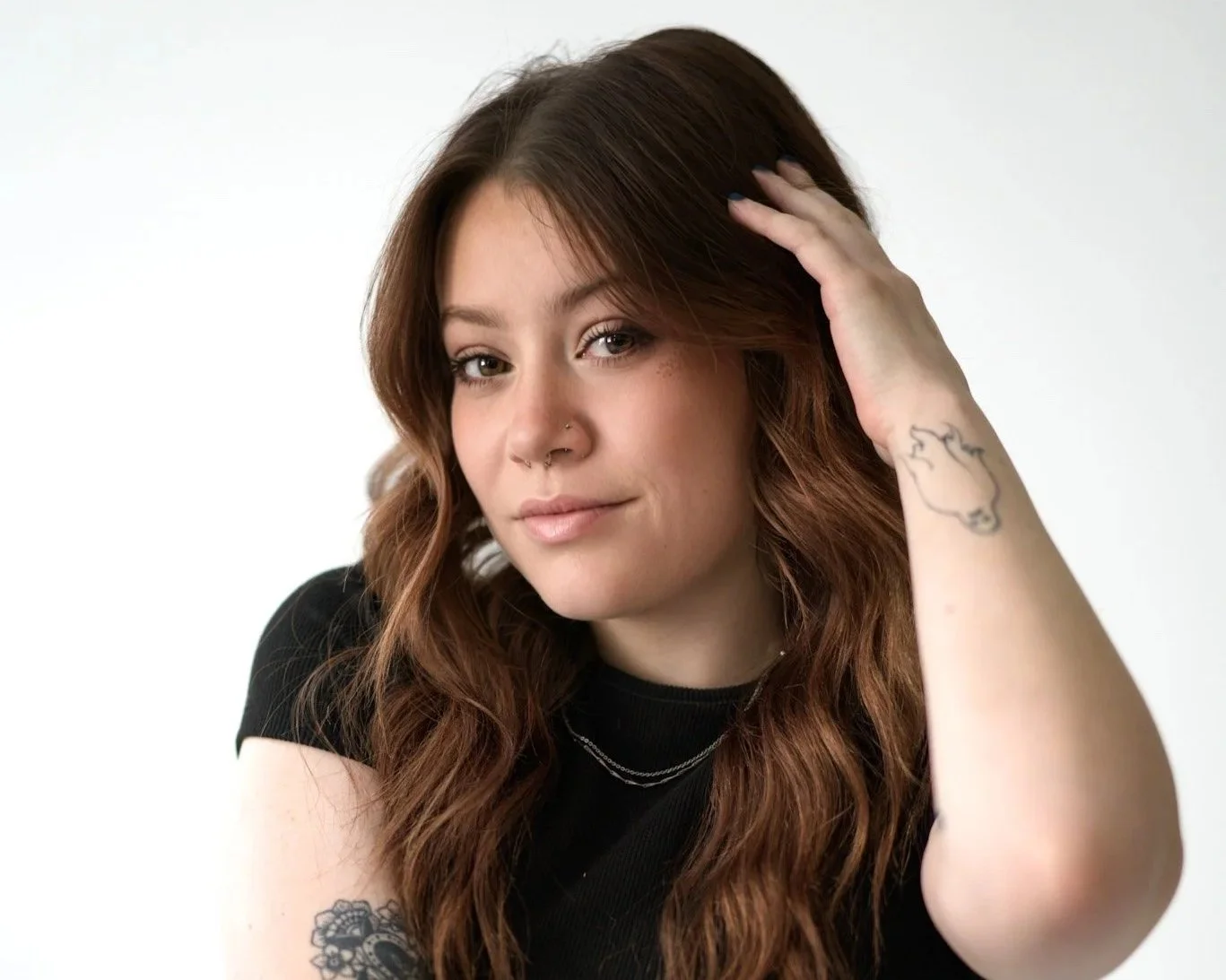 Portrait of a young woman with long brown hair, tattoos on her arm, wearing a black shirt, with a neutral expression and touching her hair, against a plain white background.