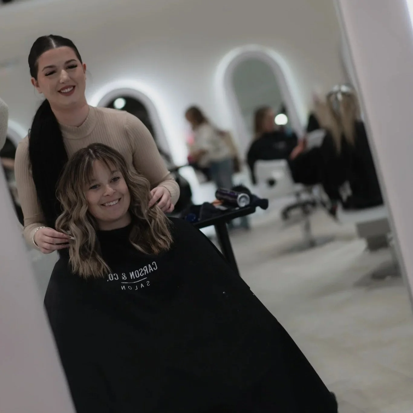 A hairstylist and a client smiling in a salon with other clients and stylists in the background.