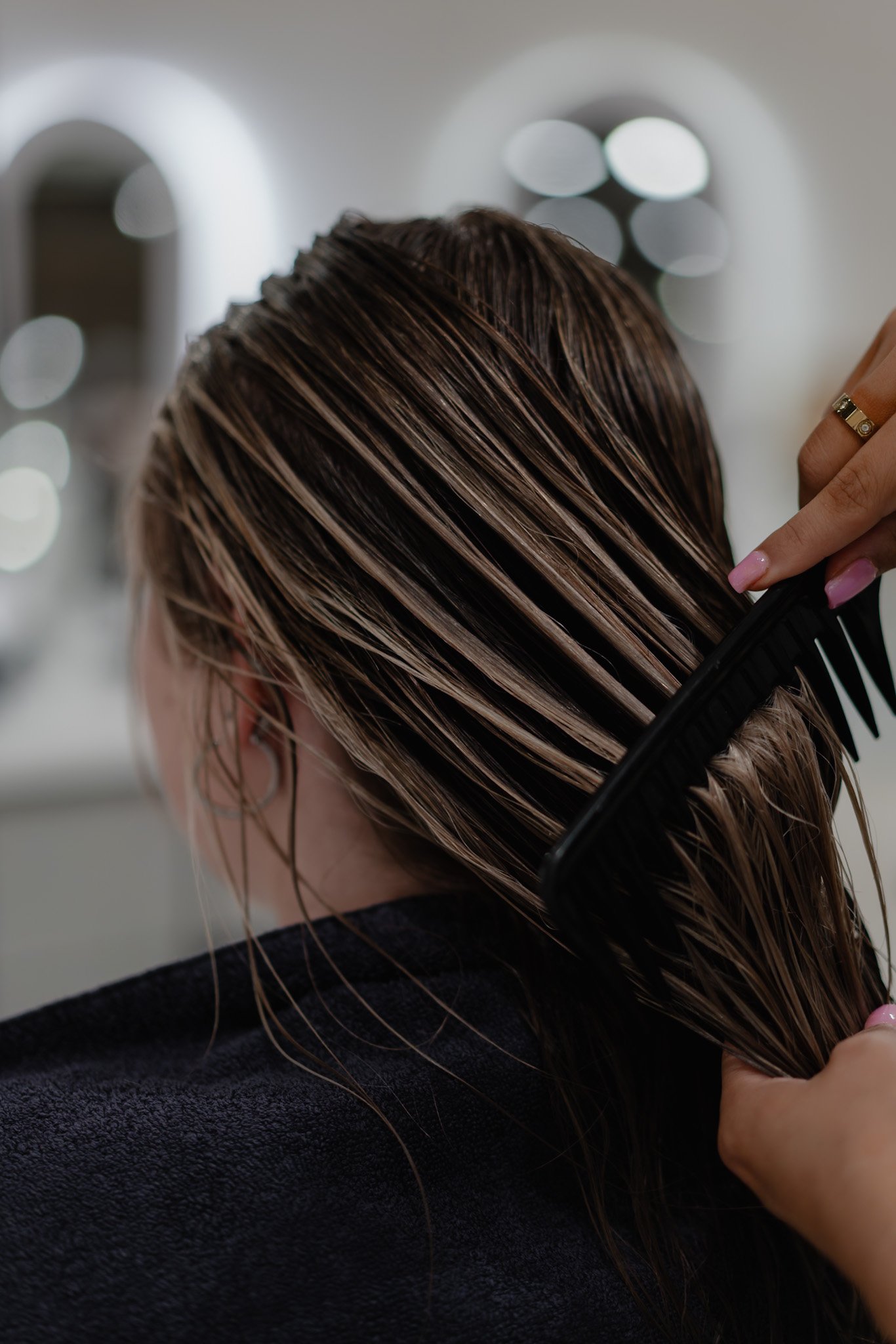 Person combing wet, light brown hair with a black wide-tooth comb in a salon setting.