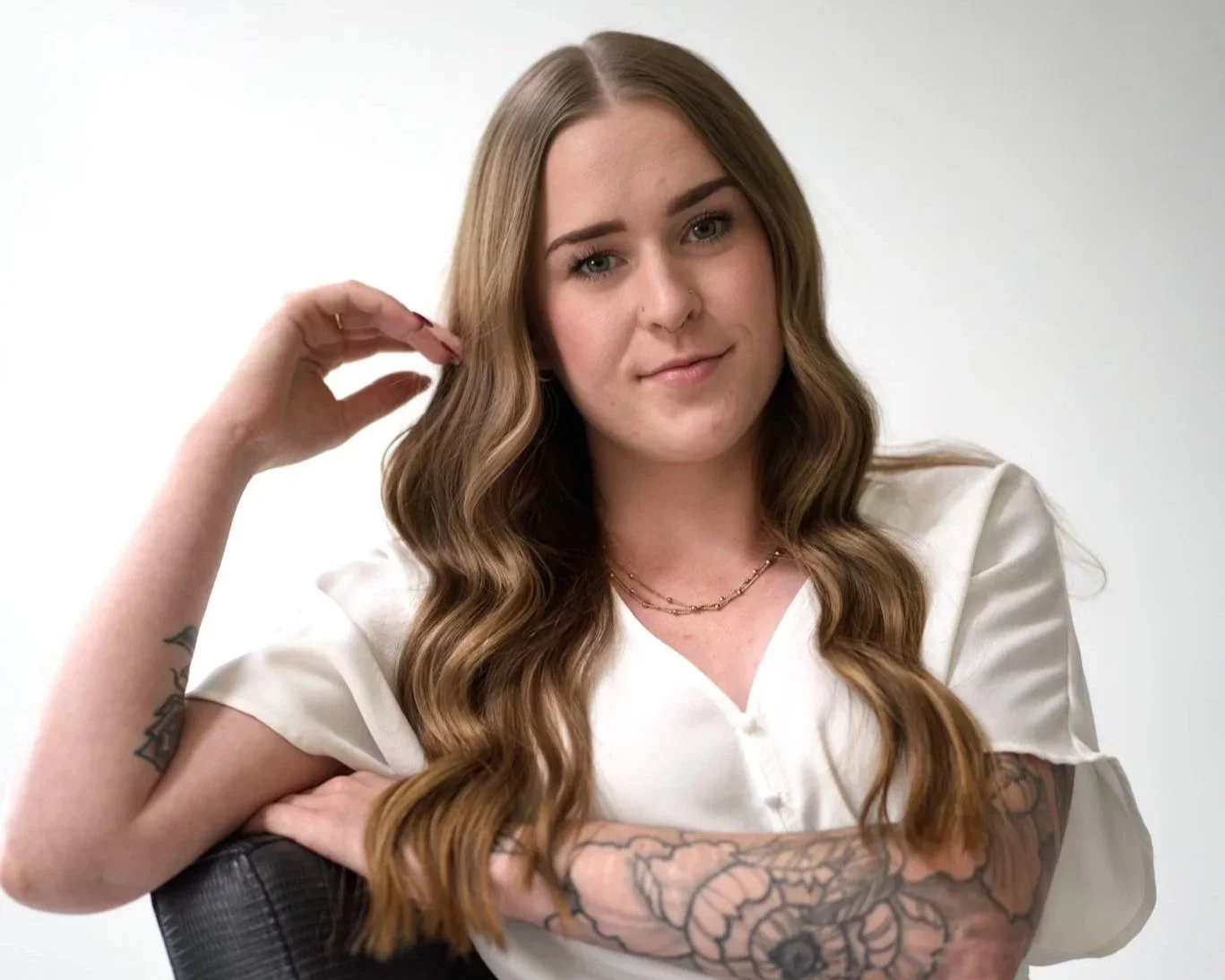 Young woman with long, wavy brown hair and tattoos on her arm, wearing a white blouse and layered necklaces, sitting on a chair with a plain background.