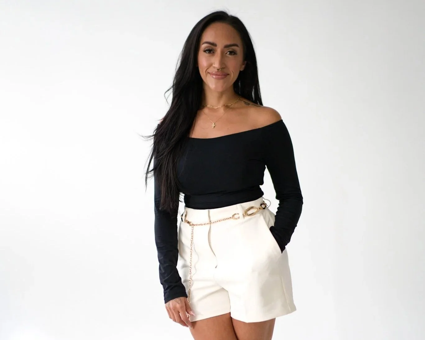 A woman with long black hair, wearing an off-the-shoulder black top and white high-waisted shorts, smiling against a plain white background.