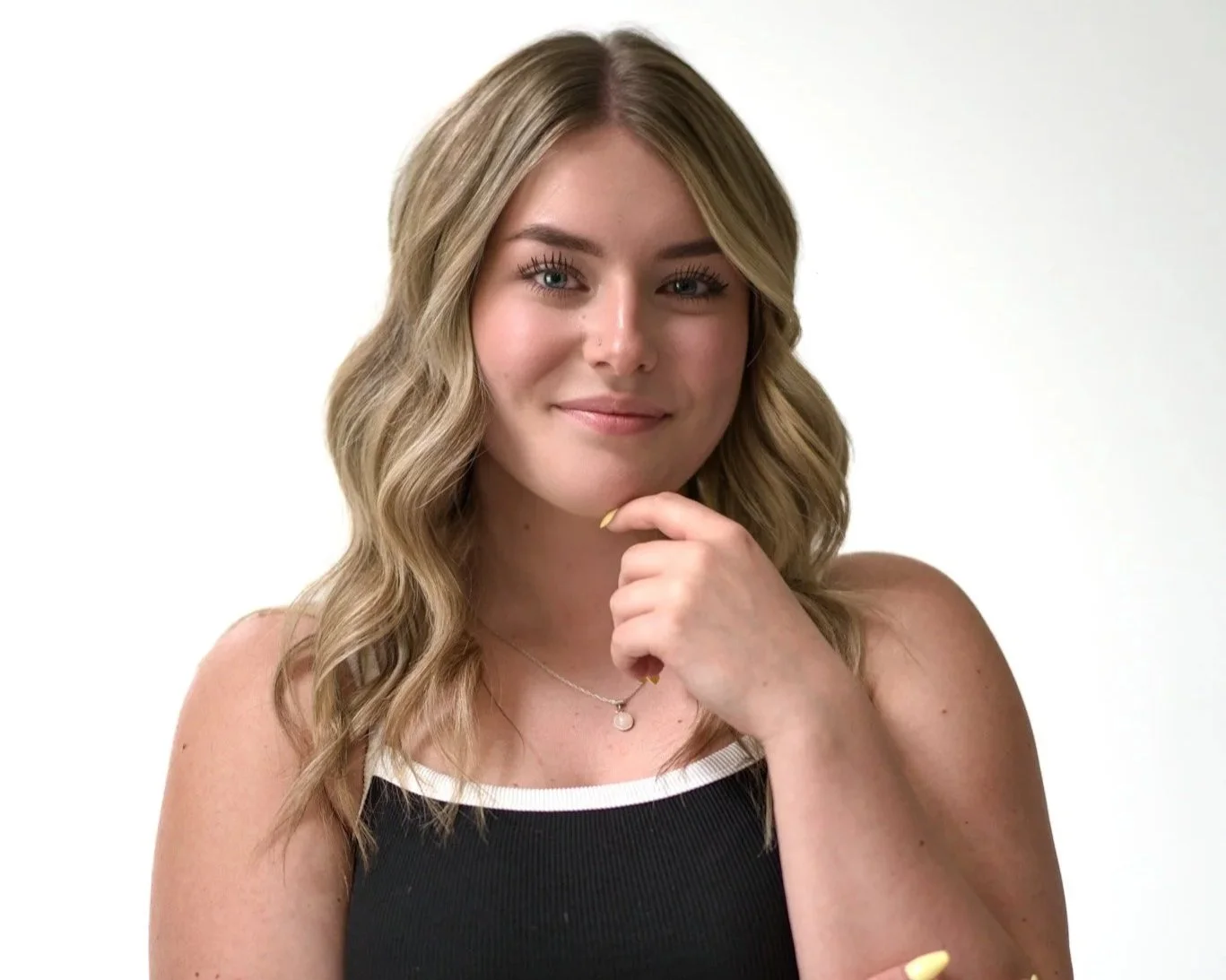 A young woman with wavy blonde hair, wearing a black tank top with white trim, is posing with her hand near her chin and smiling softly at the camera against a plain white background.