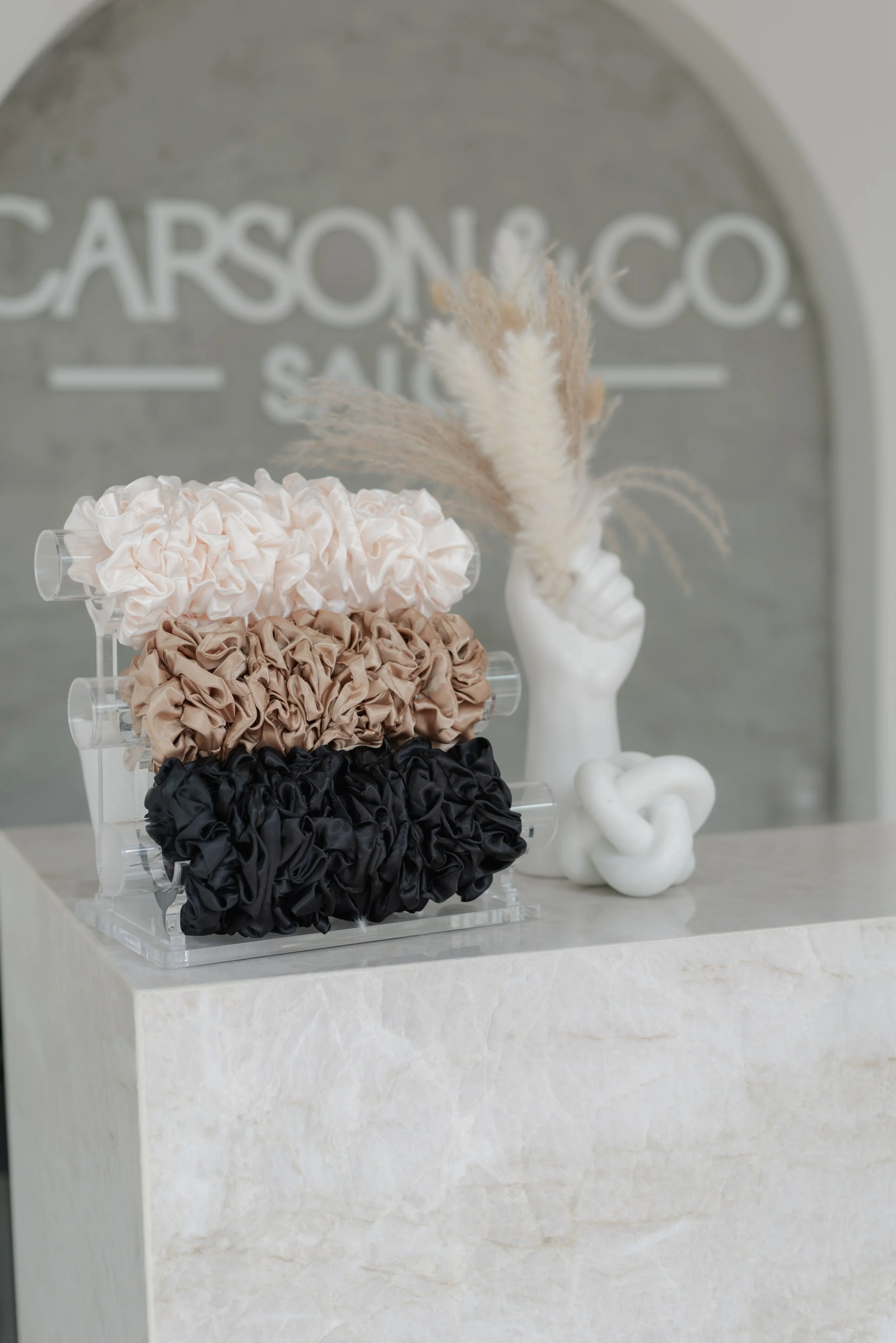 Display of fabric scrunchies in cream, beige, and black colors on a clear acrylic stand, with a white sculptural vase holding dried pampas grass behind them, in front of a gray wall with a partially visible sign reading 'CARSON & CO.'