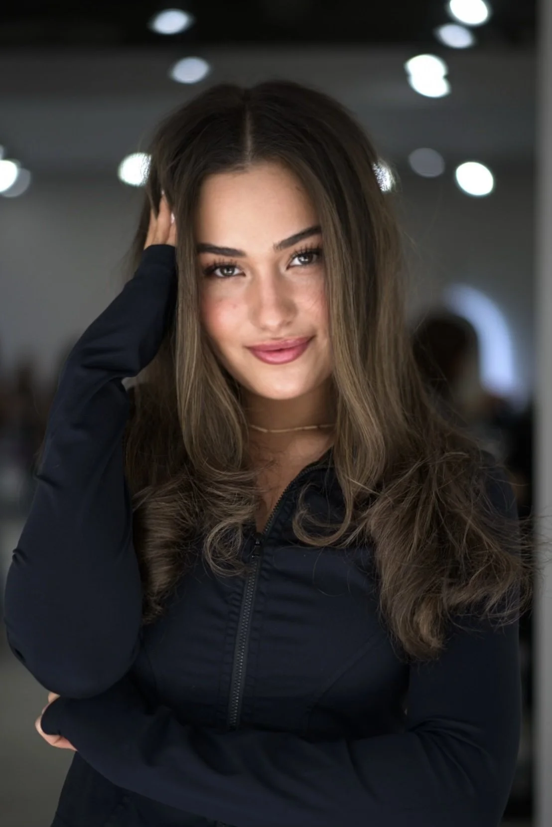 A young woman with long, wavy brown hair and light skin smiling softly, standing indoors with blurred lights in the background.