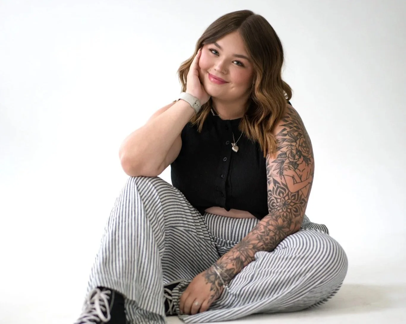A young woman with shoulder-length wavy brown hair, wearing a black sleeveless top, striped pants, and black sneakers, sitting cross-legged against a plain white background. She has a tattoo sleeve on her left arm and is smiling while resting her head on her left hand.