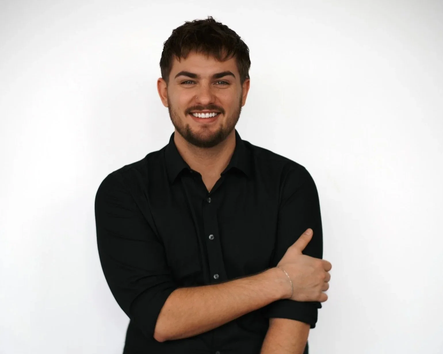 A young man with brown hair, beard, and a nose piercing smiling at the camera, dressed in a black button-up shirt, standing against a plain white background.