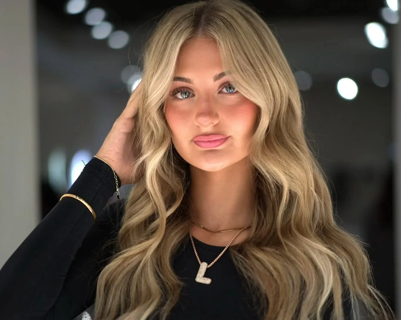 A young woman with long, wavy blonde hair, wearing a black top, and jewelry including a necklace with a letter 'L' pendant, is touching her hair with her left hand, smiling and looking at the camera, with a blurred background of lights.