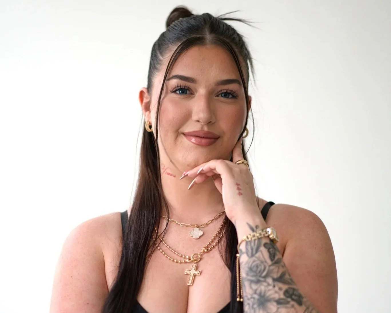 A young woman with long dark hair and blue eyes, wearing multiple gold necklaces, hoop earrings, and a black top, smiling and resting her chin on her hand, with visible tattoos on her arm and hand, against a plain white background.