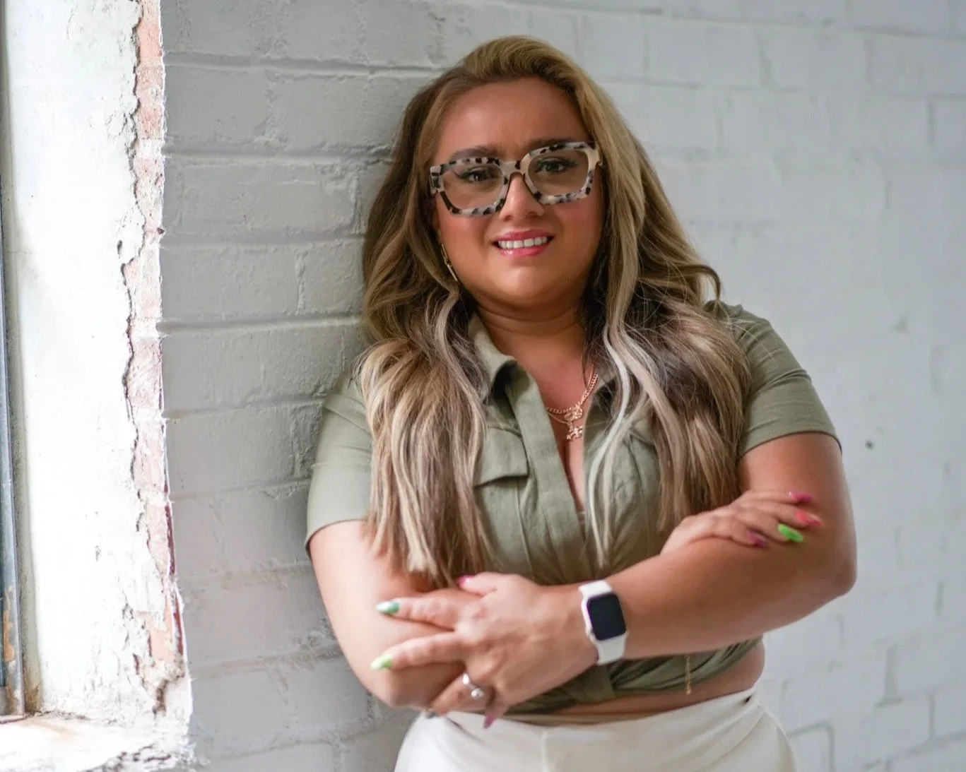 A woman standing against a white brick wall with her arms crossed, wearing glasses, a green short-sleeved shirt, and a smartwatch, smiling.