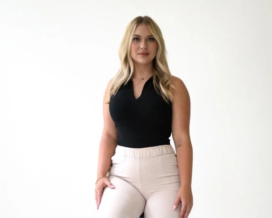 A woman with blonde hair wearing a black sleeveless top and beige pants standing against a white background.