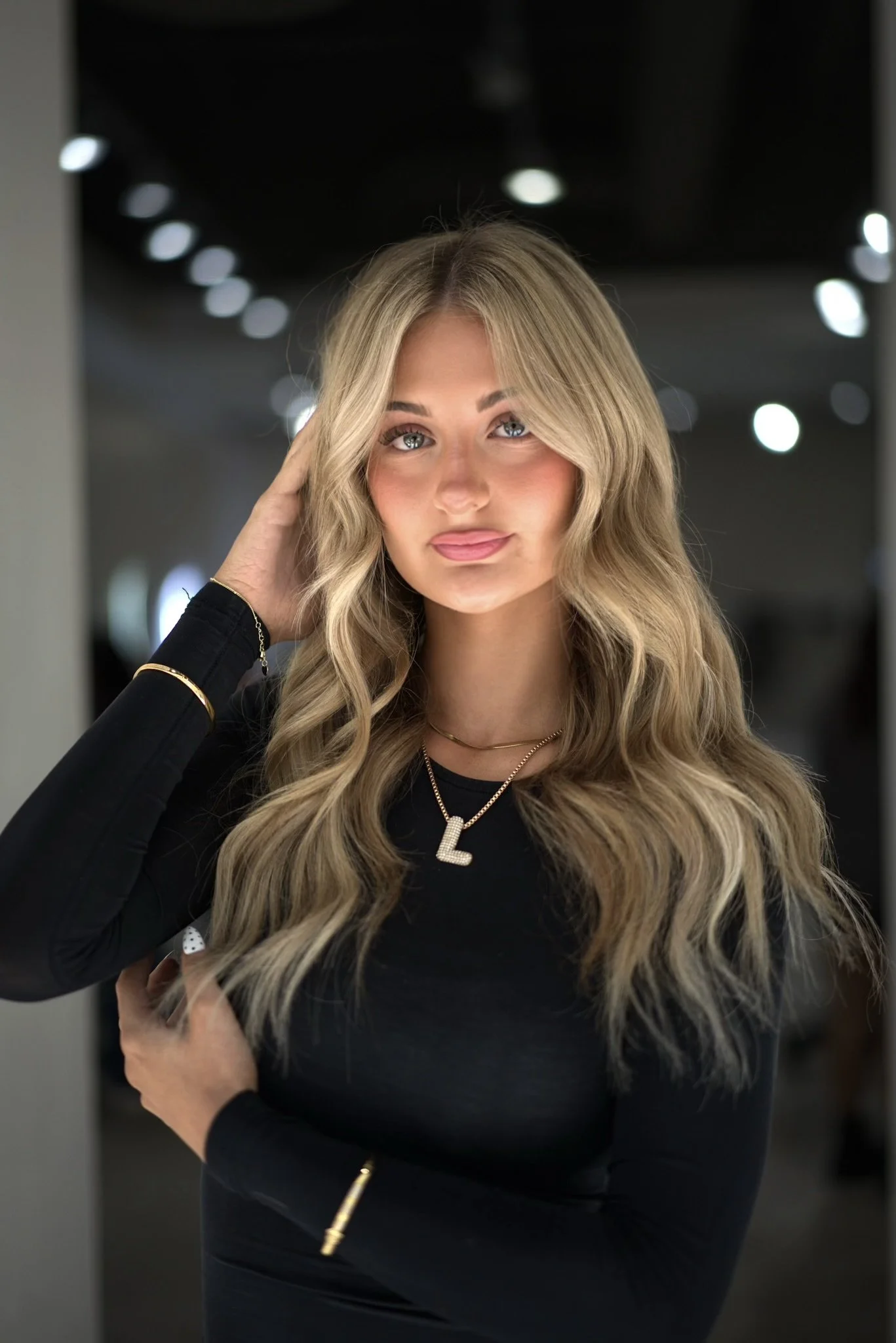 A young woman with long, wavy blonde hair and blue eyes, wearing a black top and gold jewelry, standing indoors with a blurred background of lights.