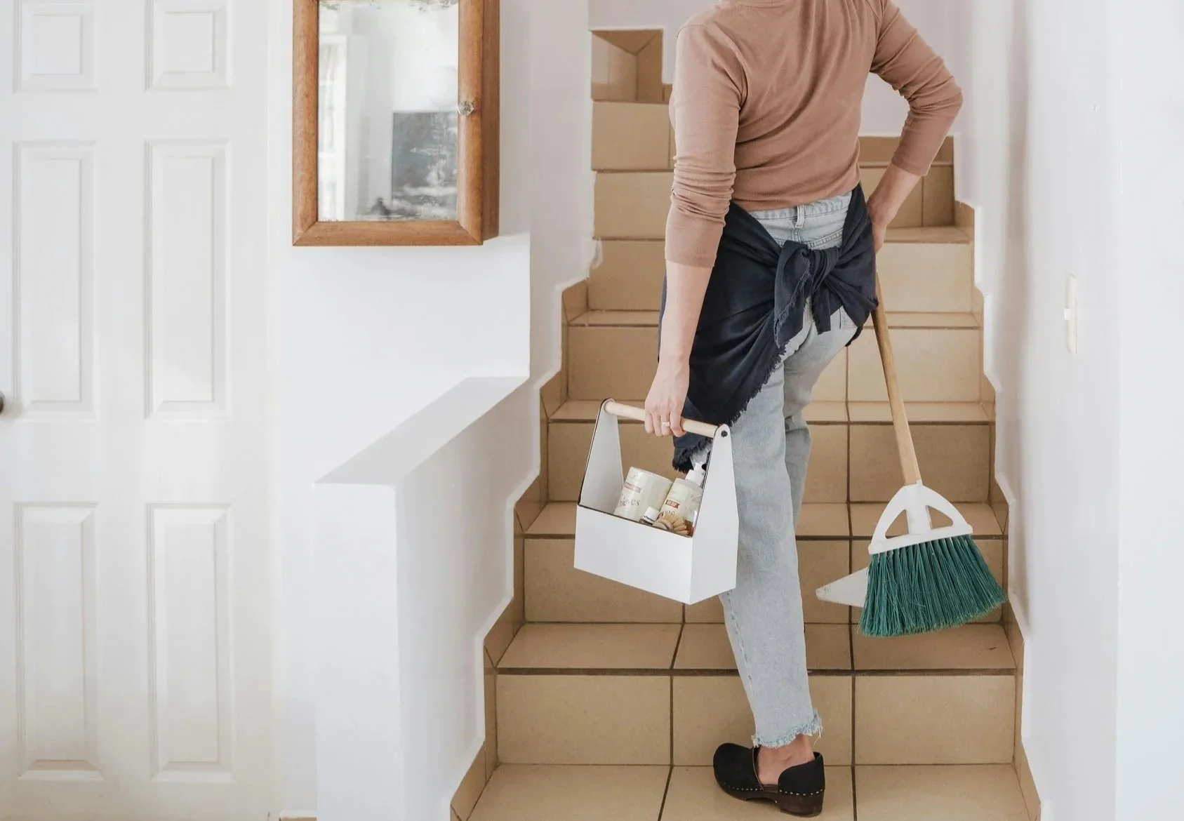 Person carrying cleaning supplies, including a box of cans and a scrub brush, while standing on a staircase.