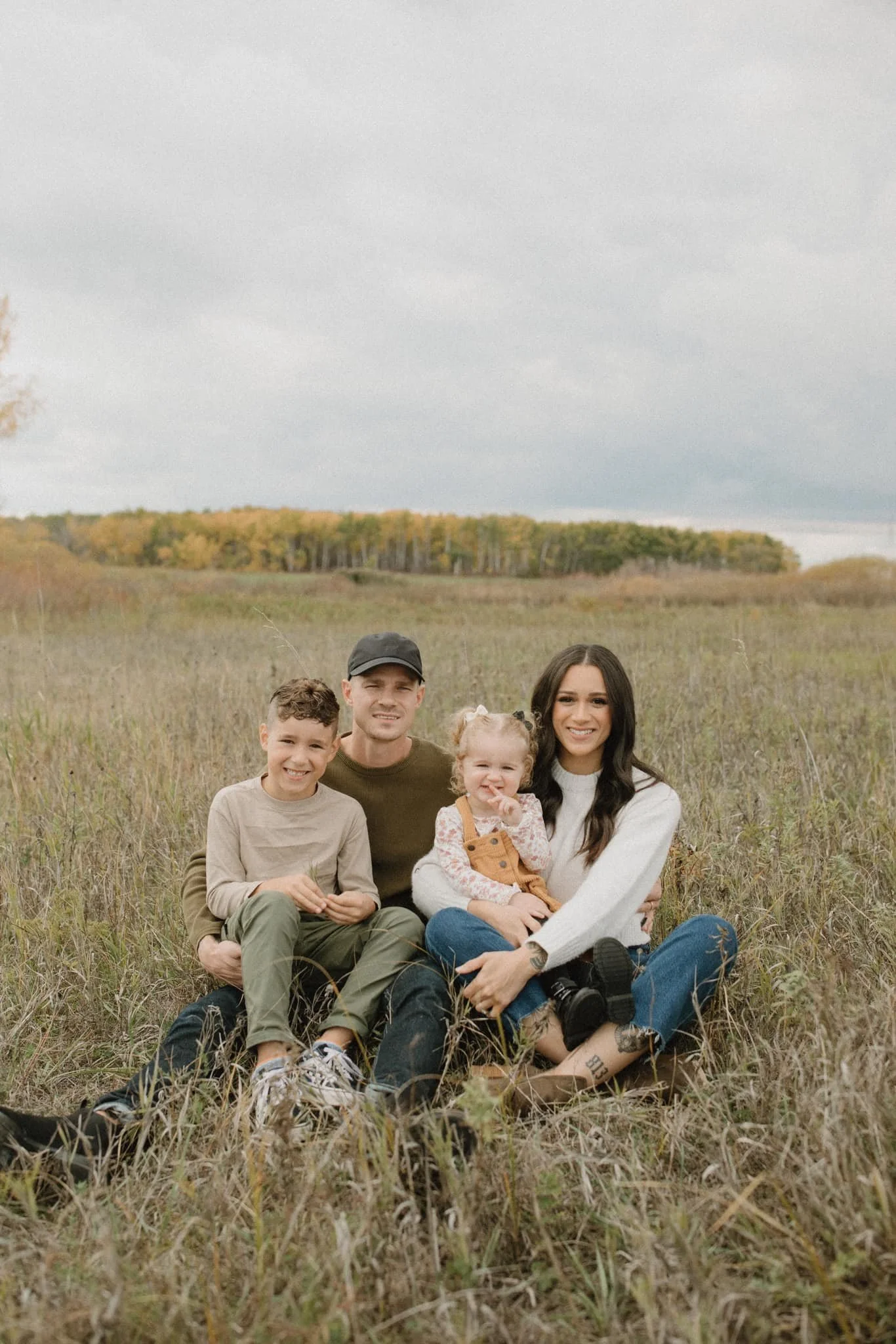 A family of four sitting on a grassy field with a blurred forest background, smiling at the camera on a cloudy day.
