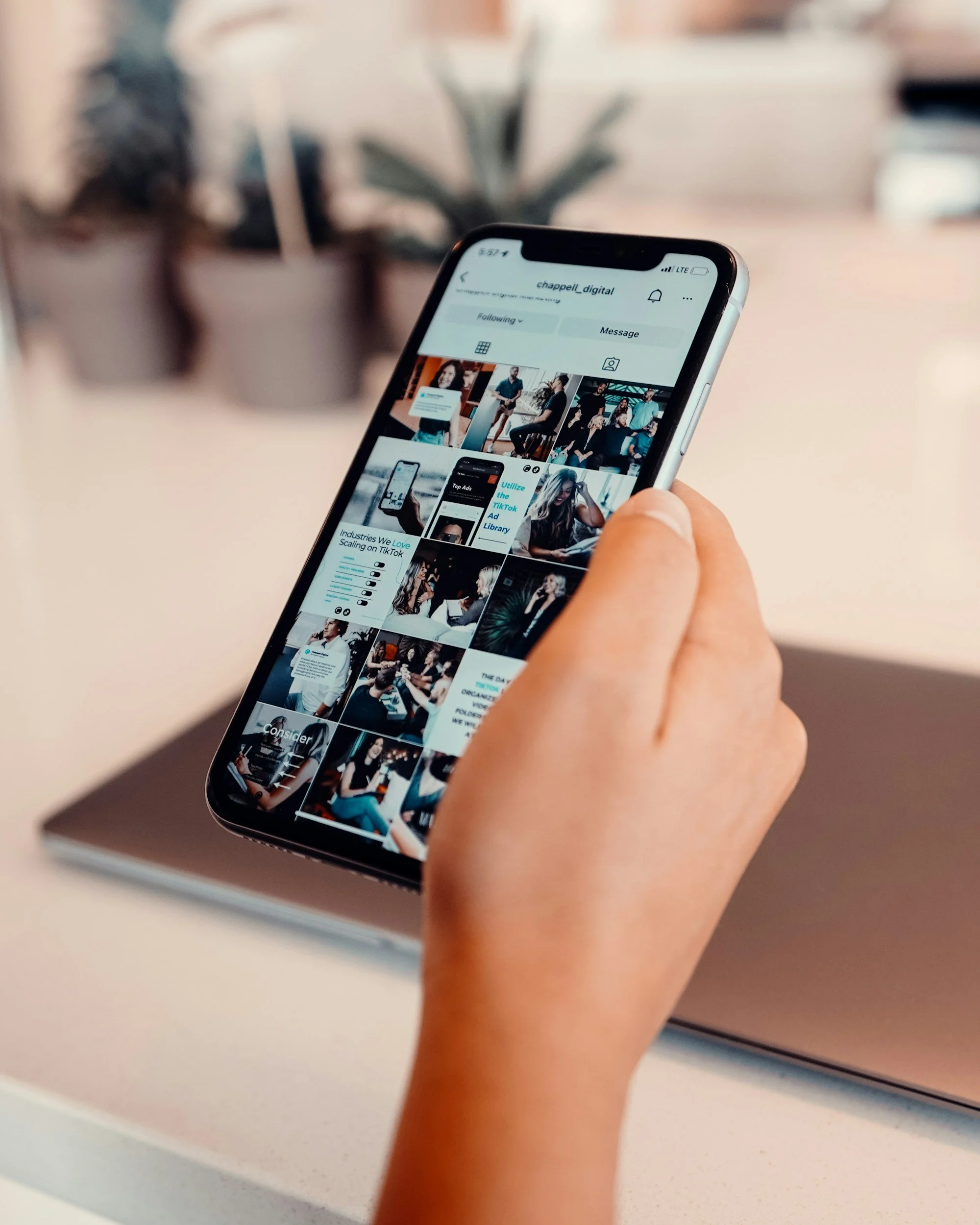 A hand holding a smartphone displaying a social media profile with various images and posts, with a background of a desk and potted plants.