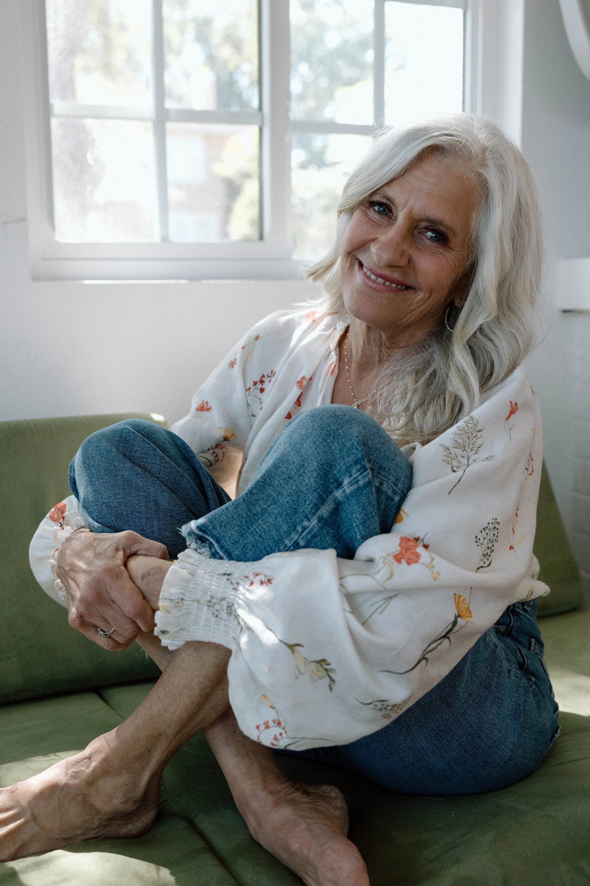 An elderly woman with long gray hair sitting on a green couch, smiling, with her knees pulled up to her chest, in front of a window with natural light.
