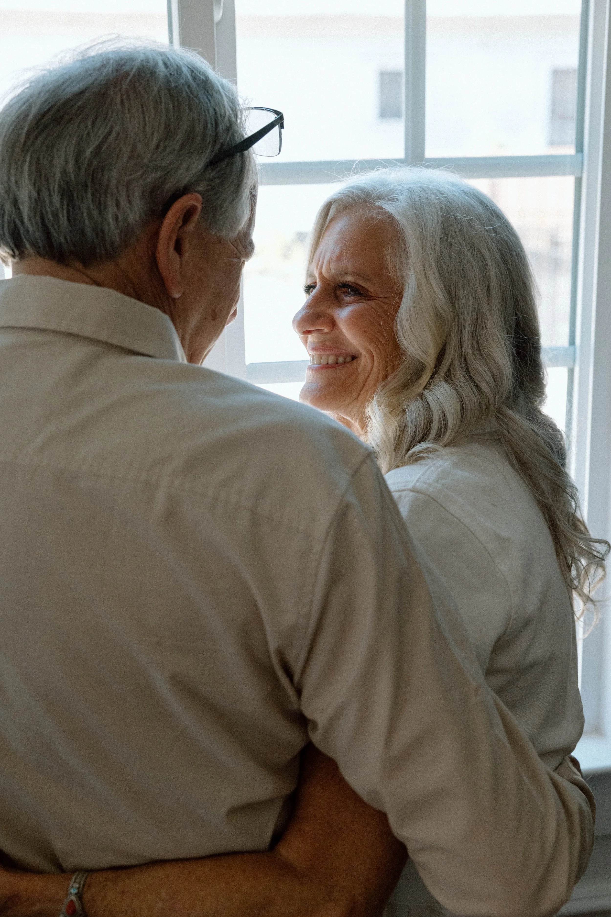 An elderly couple smiling at each other near a window.