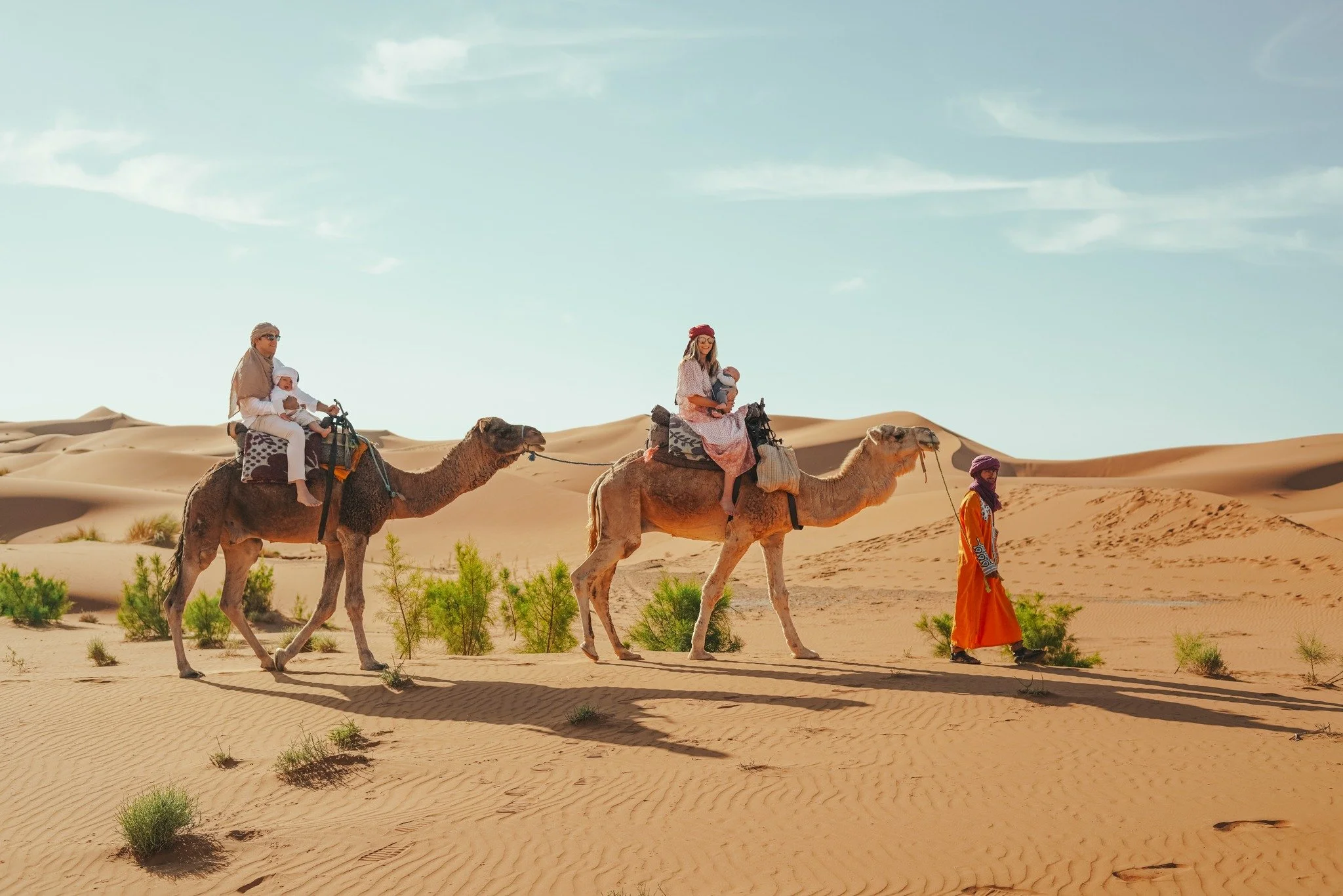 People riding camels across desert sand dunes, with an orange-clad guide walking alongside.
