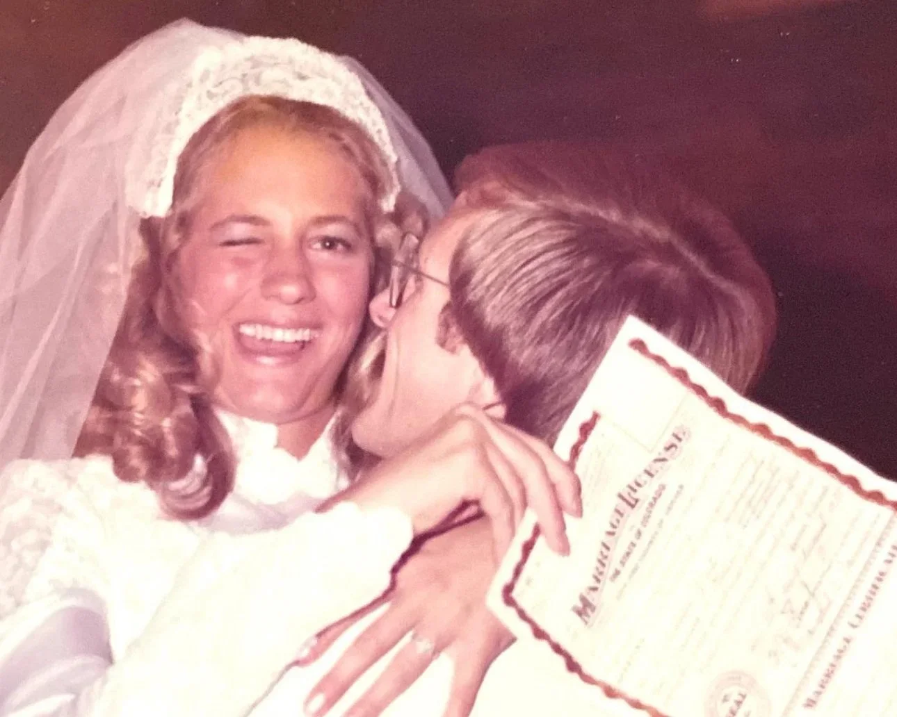 A wedding photo of a bride and groom sharing a kiss, with the groom holding a marriage license in front of them.