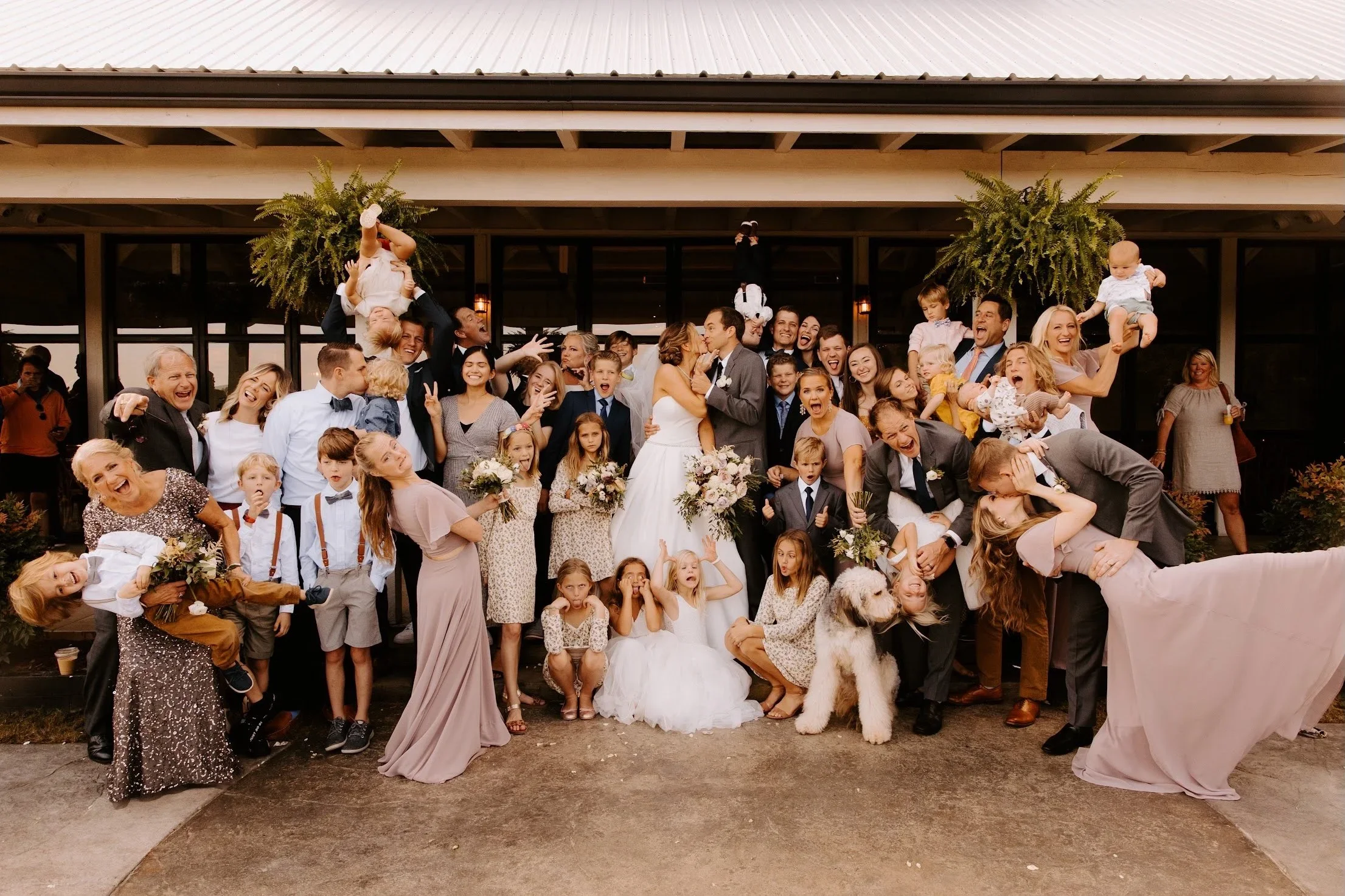 Group photo of a wedding celebration with the bride and groom kissing, surrounded by friends and family, some holding children, with two large hanging plants in the background.