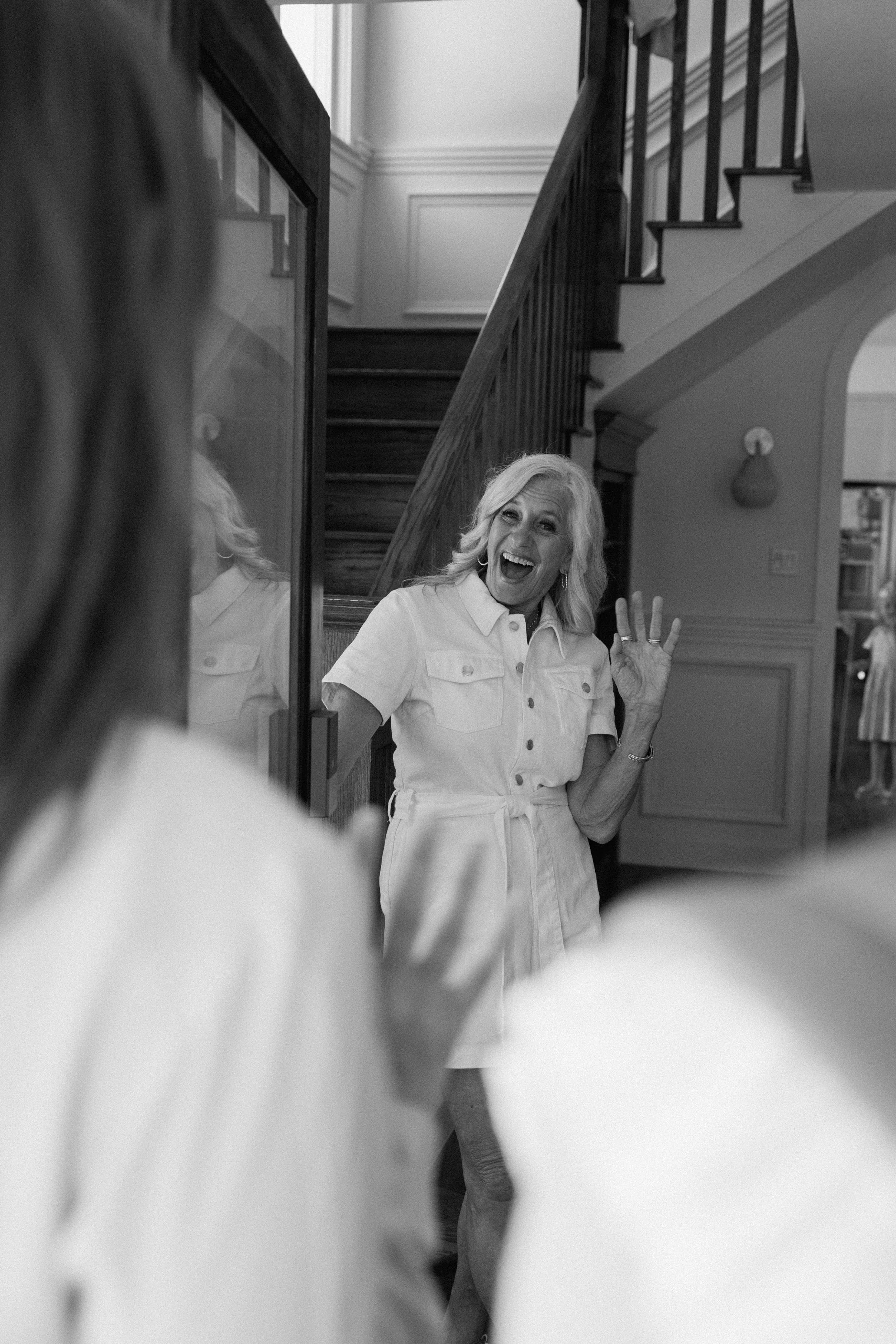 An elderly woman with blonde hair smiling and waving at someone while standing near a mirror inside a house.