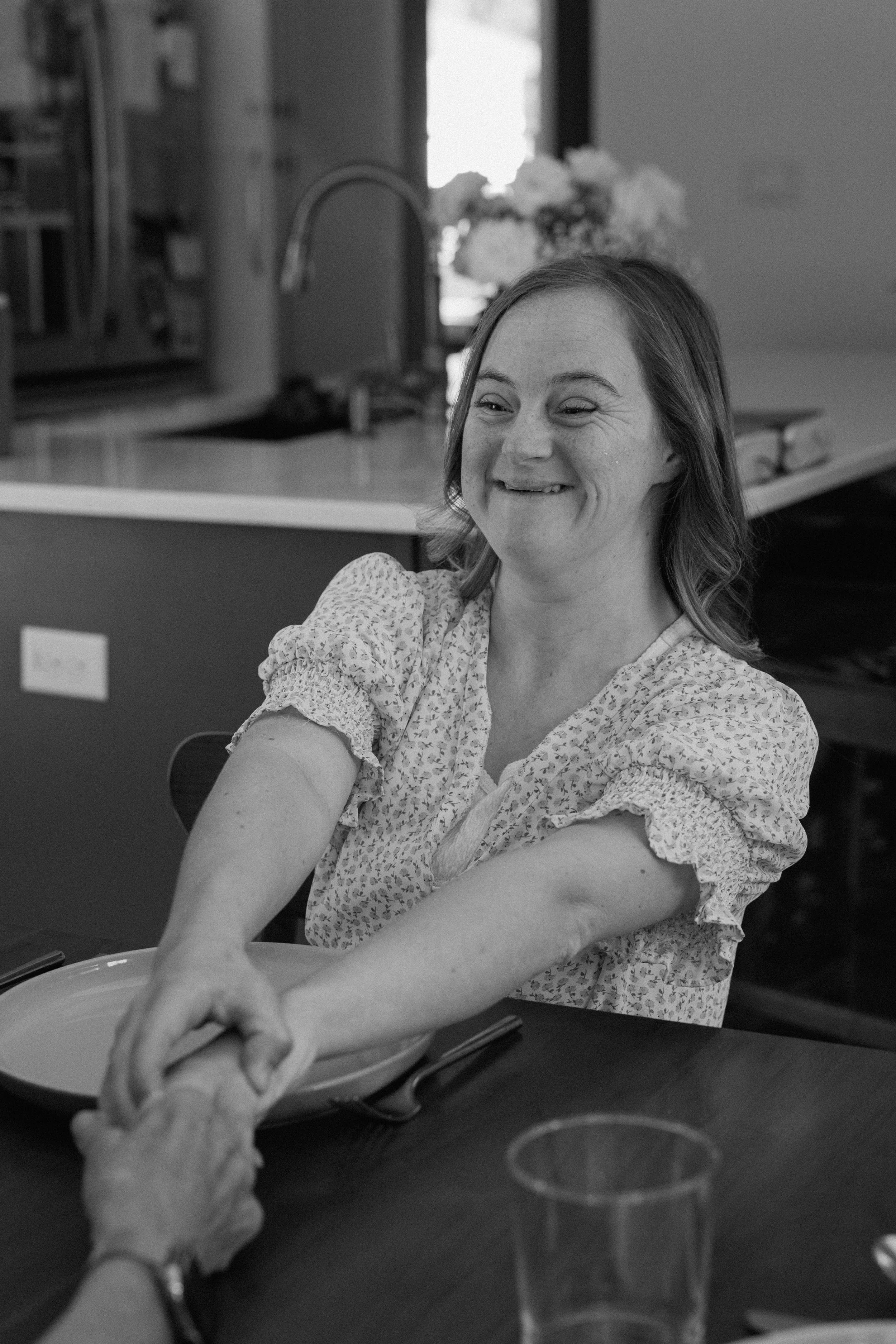 A woman smiling and reaching across a table, holding hands with someone else in a dining setting.