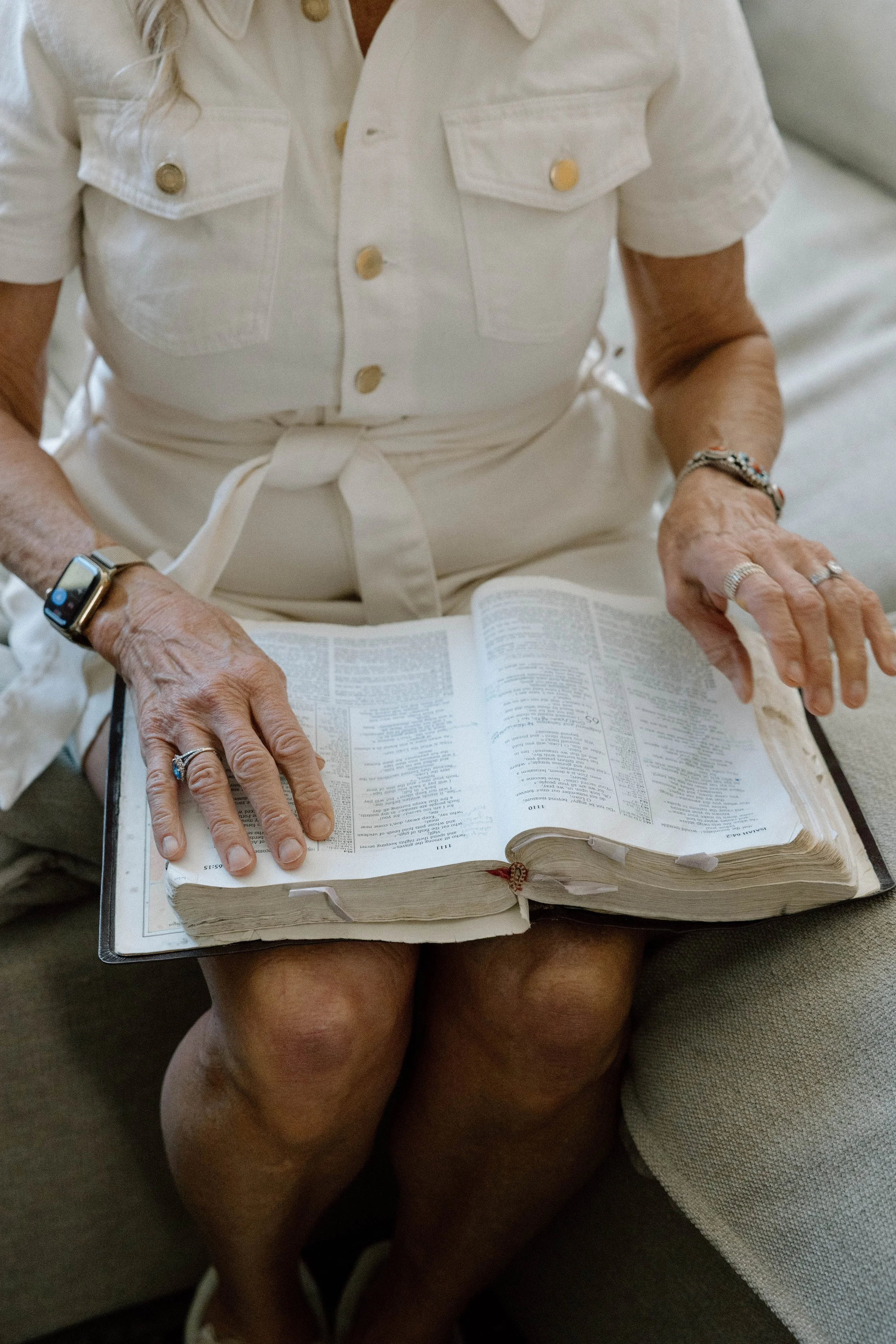 An elderly woman is sitting on a couch reading a large Bible or dictionary. She is wearing a white dress with short sleeves and has various rings and a watch on her hands.