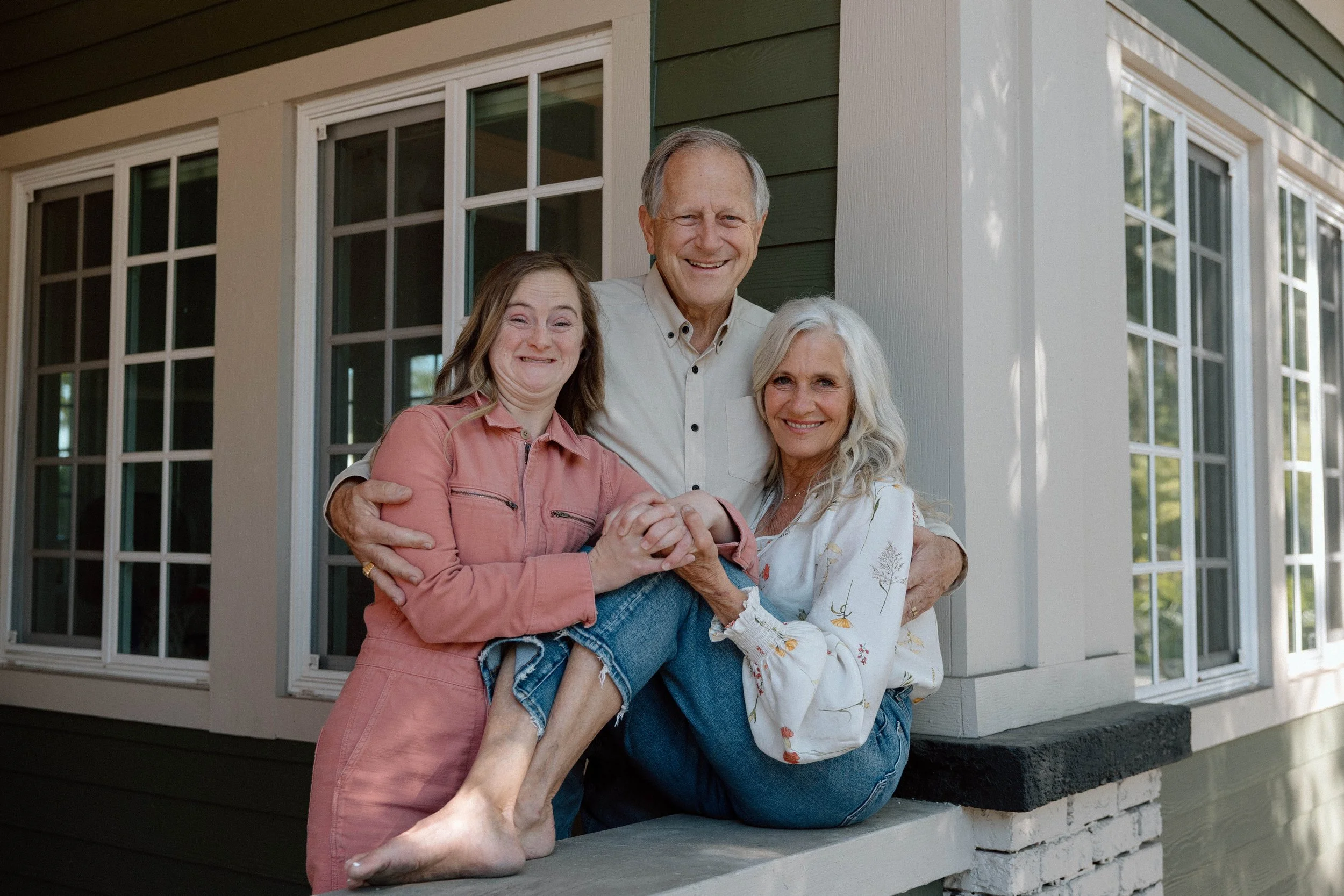 A happy family of three standing outside a house, with a woman sitting on the porch ledge, supported by her husband and daughter, all smiling at the camera.