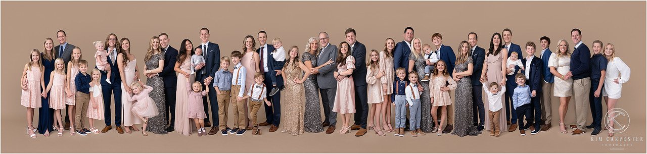 Large family portrait with multiple generations and diverse family members, including adults, teenagers, and children, standing together against a plain beige background.