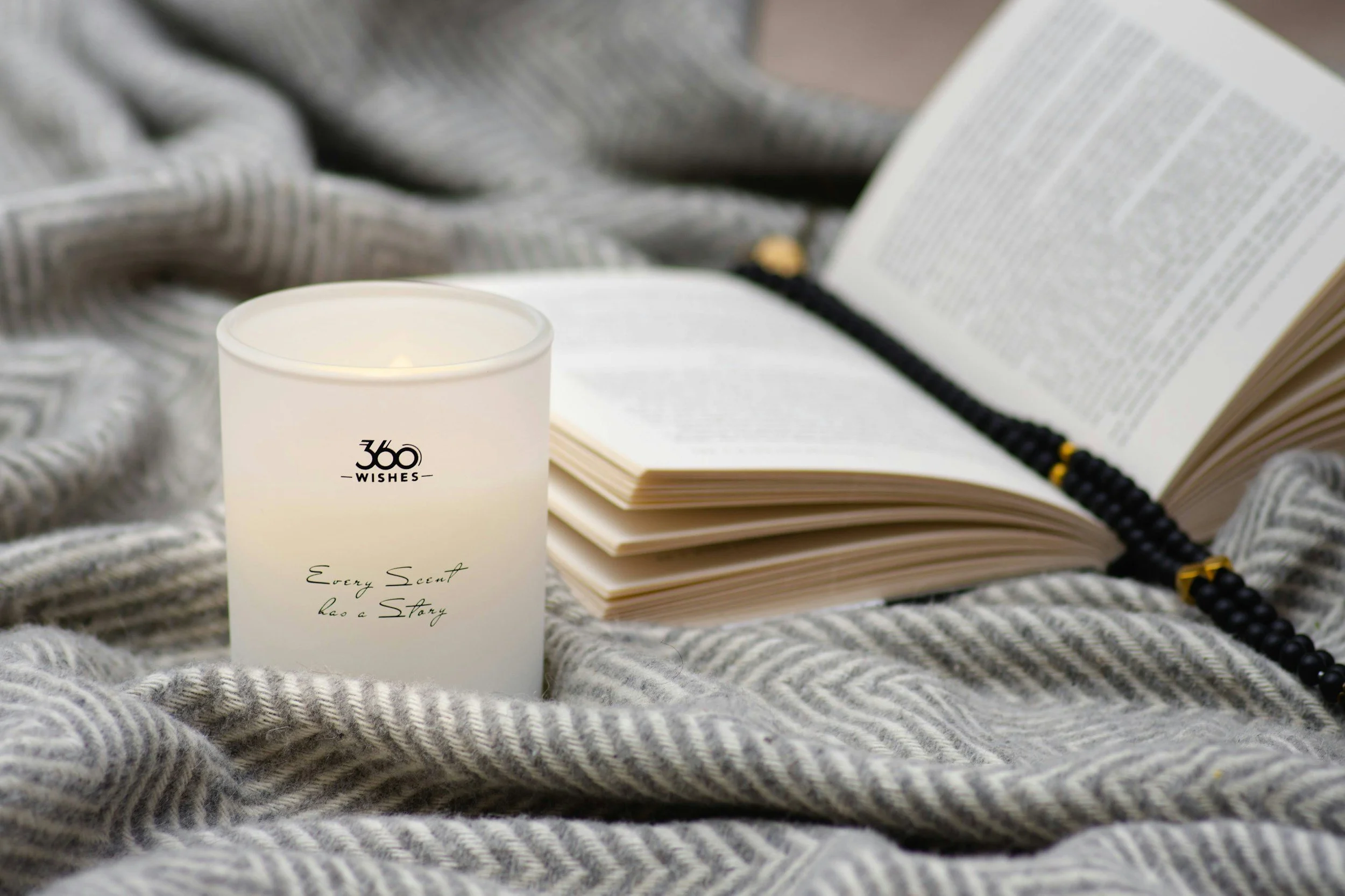 A white candle with black text reading "360 Wishes" and "Every Scent Has a Story" sits on a gray and white patterned fabric, with an open book and a black beaded rosary with gold accents lying beside it.