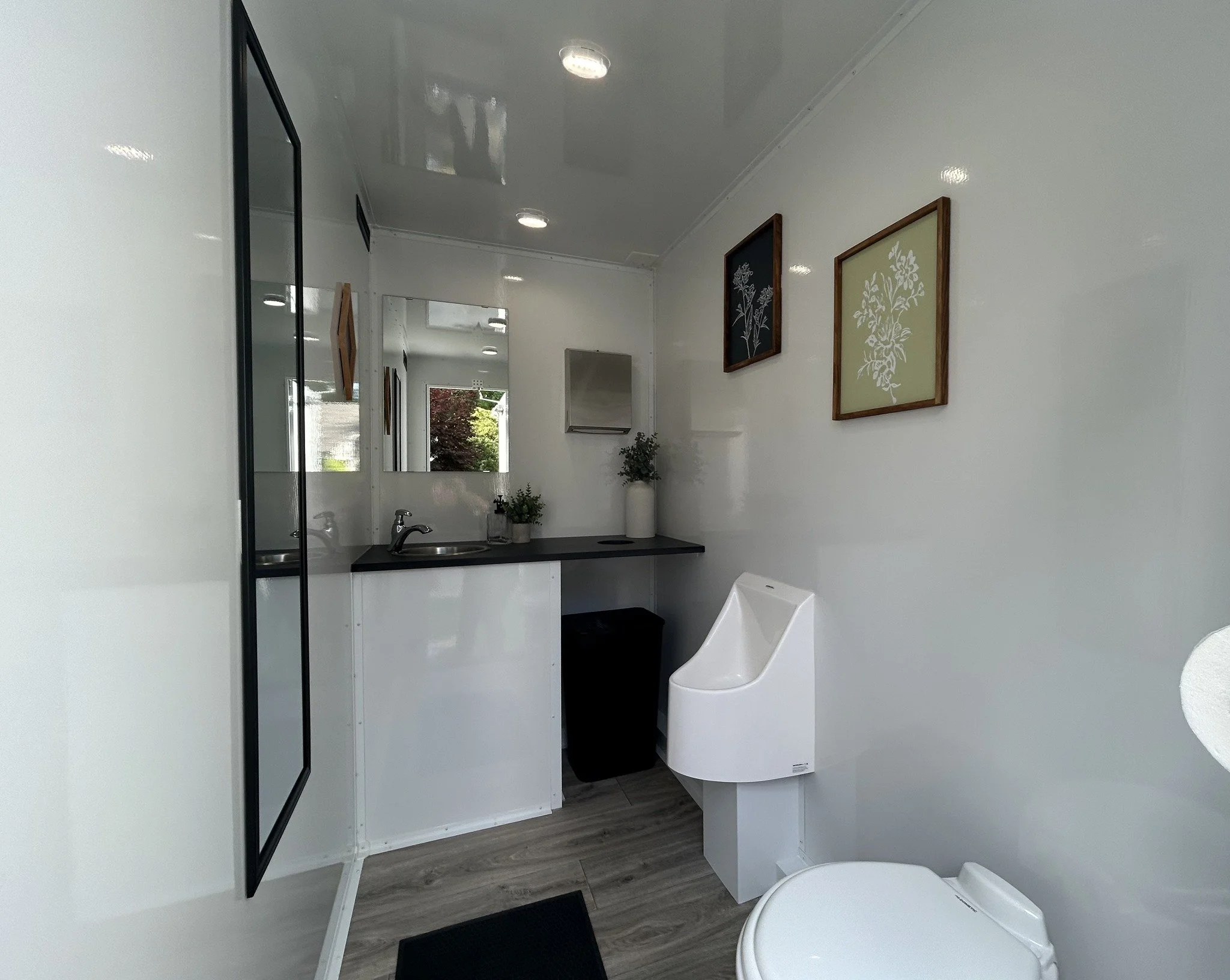A small, clean bathroom with white walls, a toilet with a built-in sink on top, a black countertop with a sink and faucet, a mirror, and minimalist framed artwork on the wall. There are green plants near the sink and a window showing outdoor greenery.