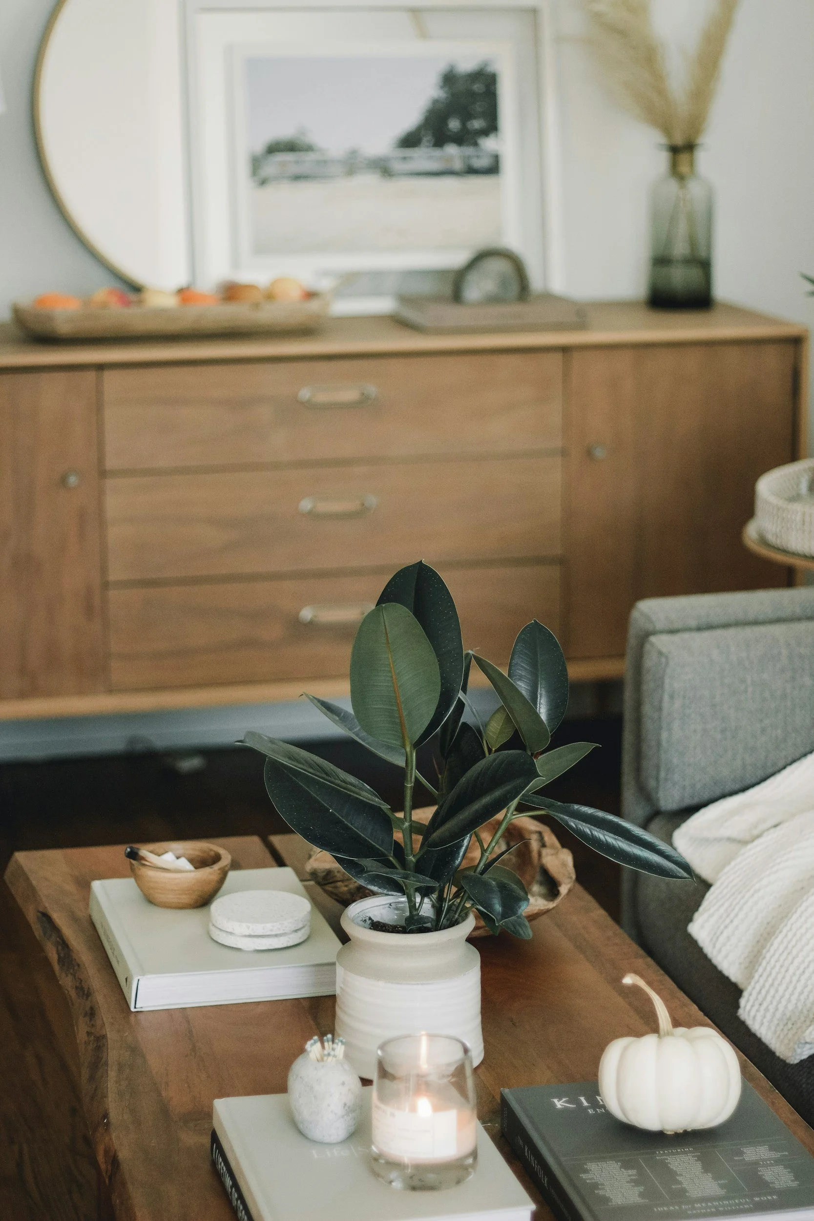 A cozy living room with a wooden coffee table decorated with a white pumpkin, a lit candle, a potted plant, books, and small decorative items. In the background, there is a wooden sideboard with a tray of oranges, a framed picture, and a tall vase with dried plants.