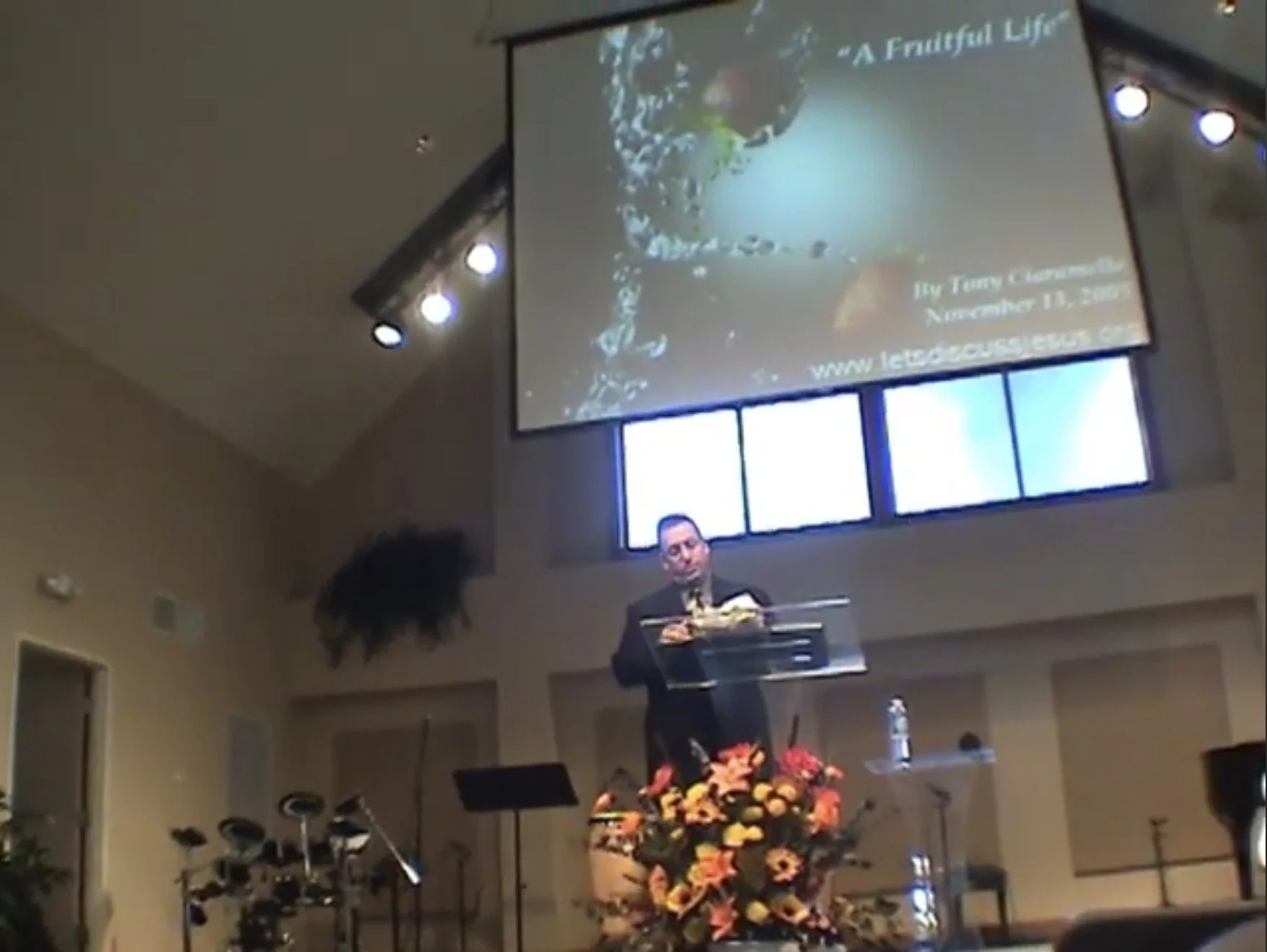 A man standing at a transparent podium giving a presentation or speech in a church or auditorium with a large projection screen behind him. The screen displays the title 'A Fruitful Life' and some additional text, including his name, date, and website. There are flowers in front of the podium, and musical instruments like drums are visible on the left side of the stage.