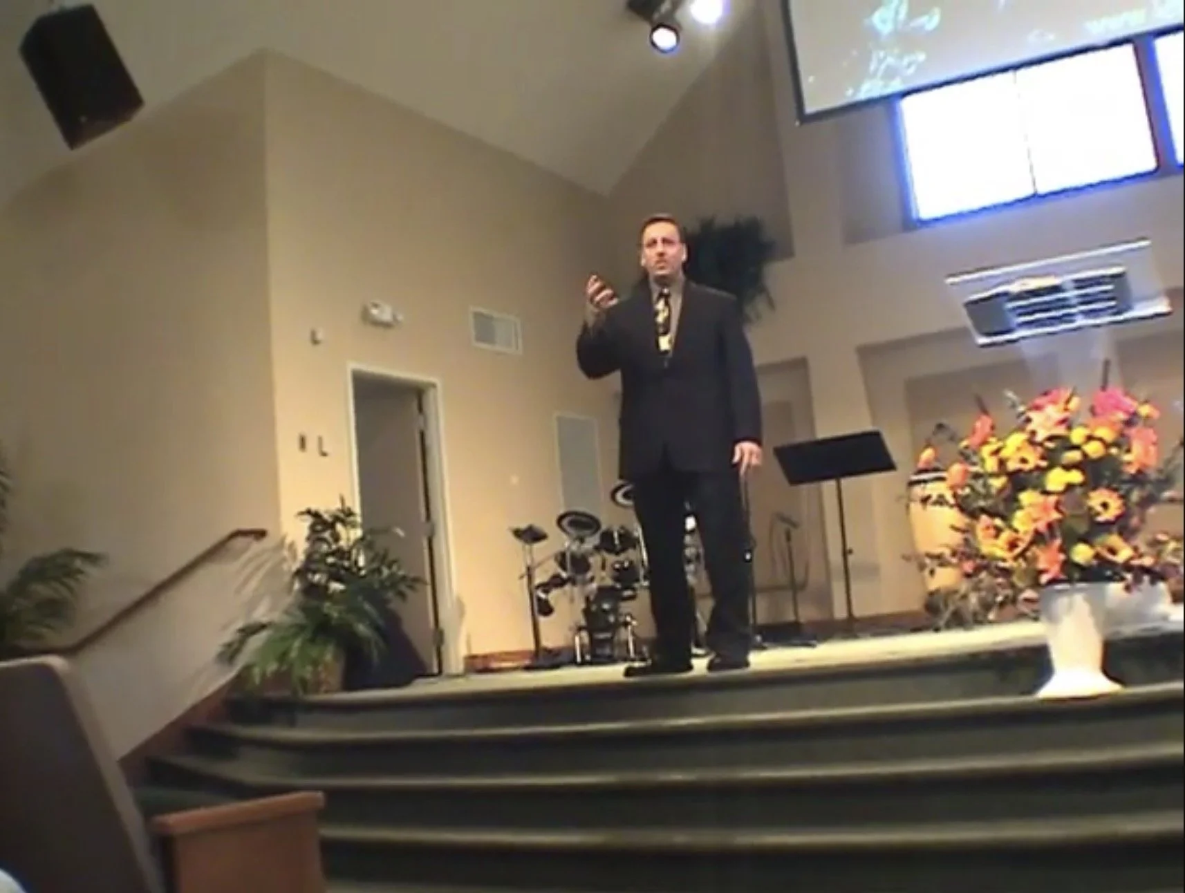 A man in a dark suit and patterned tie standing on a church stage holding a microphone, with musical instruments and colorful flower arrangements around him.