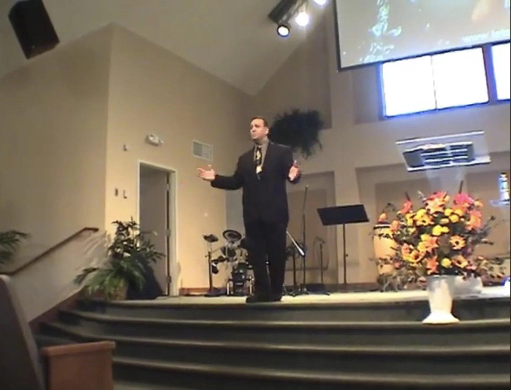 A man in a black suit and tie speaking on a stage decorated with flowers, with musical equipment and plants in the background.