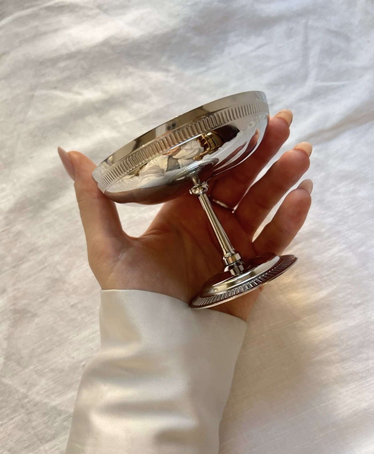 A person holding a small silver trophy with a rounded, ridged top and a short stem, with a beige satin or silk fabric background.