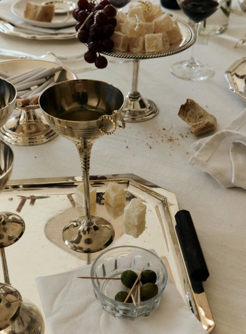 A table setting with silver and gold serving dishes, a bowl of green olives with toothpicks, a glass of red wine, a dish of bread cubes, and a small chunk of cheese and bread crumbs.