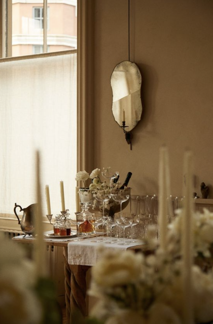A cozy dining or event space with a table decorated with flowers, candles, and glassware, near a window with beige curtains, and a vintage mirror on the wall.