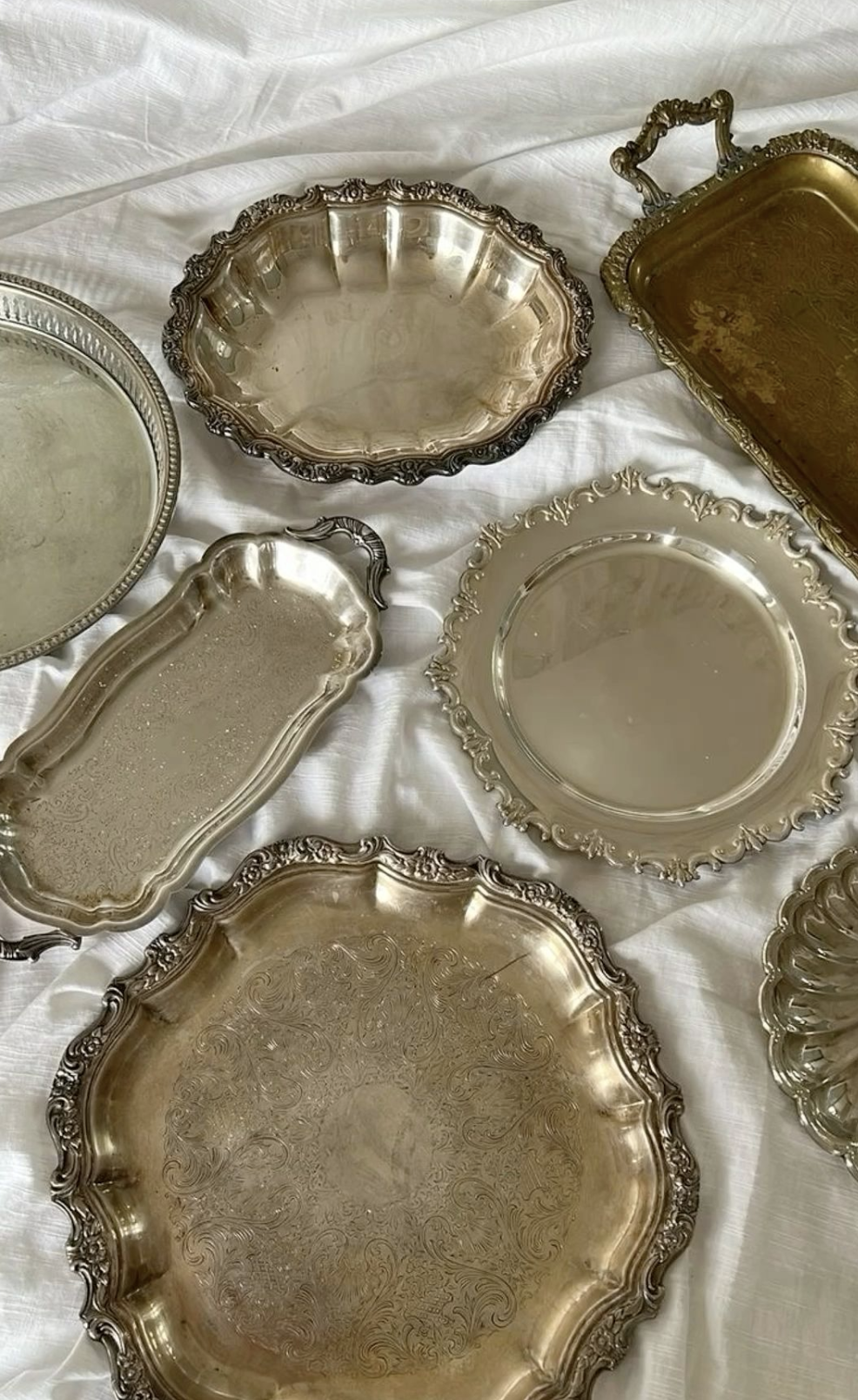 Collection of vintage silver trays and serving dishes with ornate edges on a white cloth background.