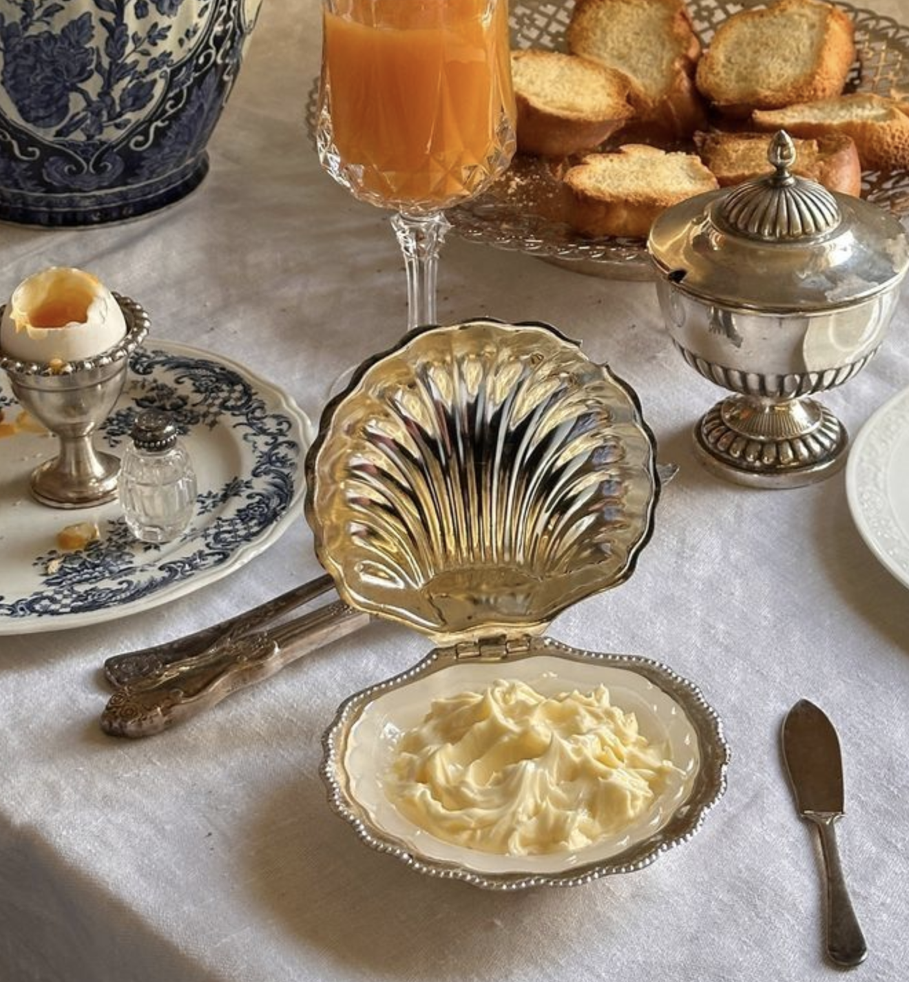 An ornate silver butter dish with butter, a plate of sliced bread, a glass of orange juice, and a dollop of butter in a ceramic dish, on a white tablecloth.