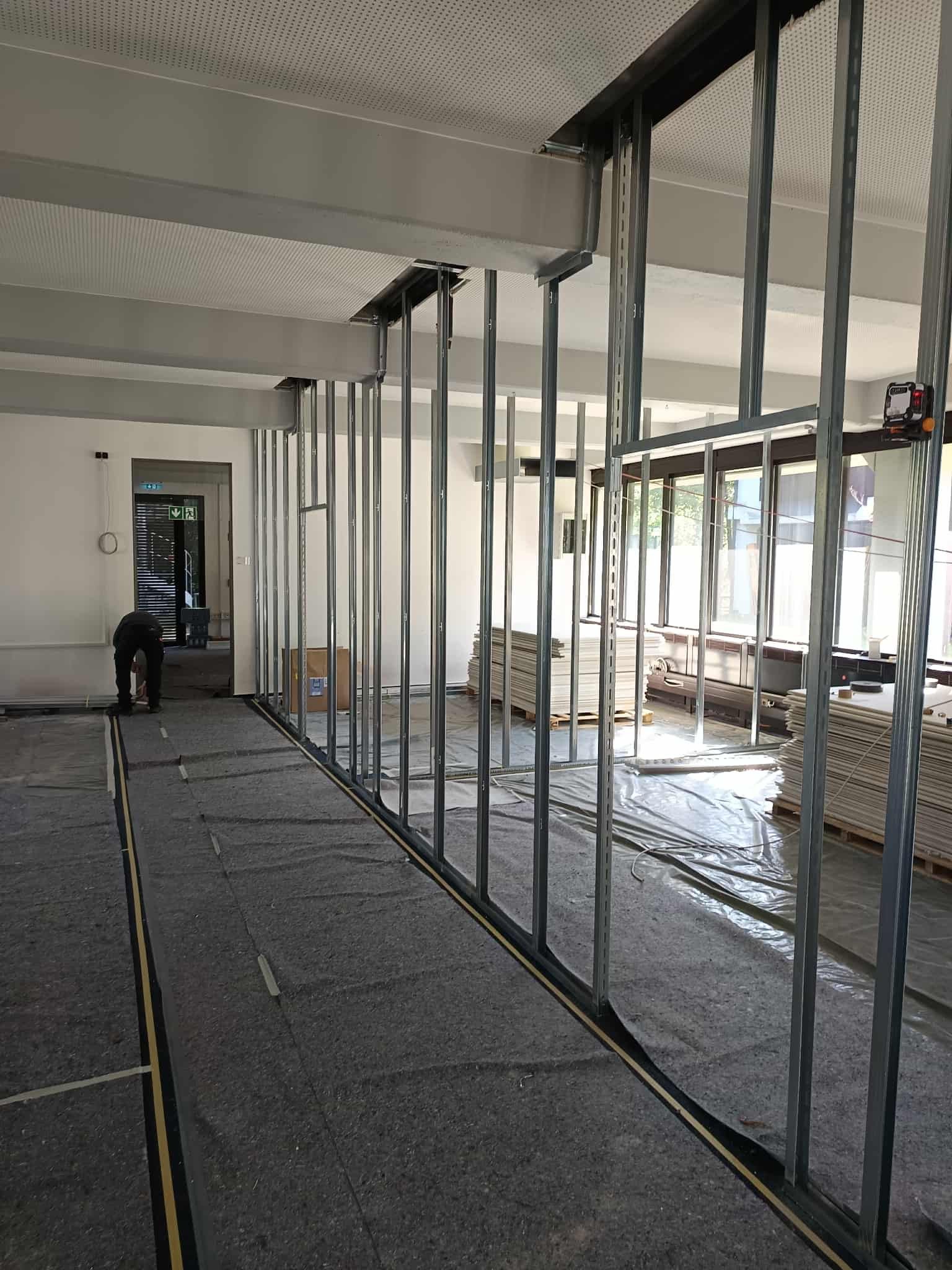 Bauarbeiten im Innenraum mit Metallrahmen für Wände und Bauzubehör, Arbeitsmaterialien und Fenster in einem Raum, vermutlich im Umbau oder Neubau.