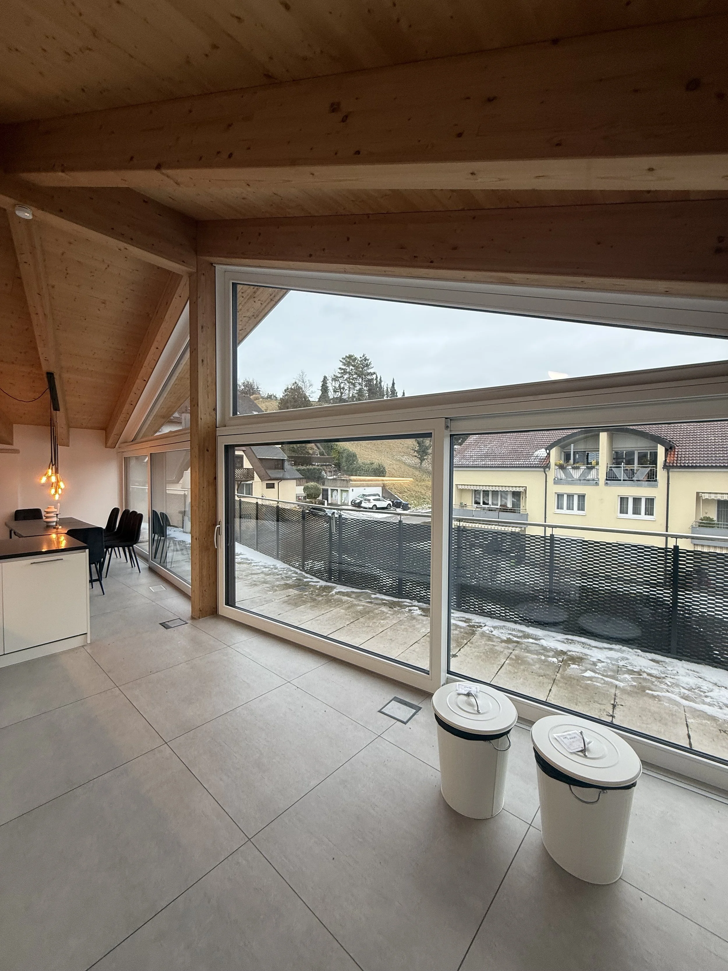 Modernes Wohnzimmer mit großen Fenstern, Blick auf den Balkon und Nachbarhäuser, Holzdecken und Fliesenboden.